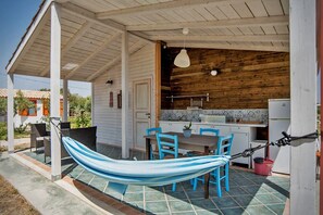 Un espace extérieur abrité comprenant un hamac, des chaises bleues, une table et un coin cuisine avec des murs en bois et un sol carrelé.