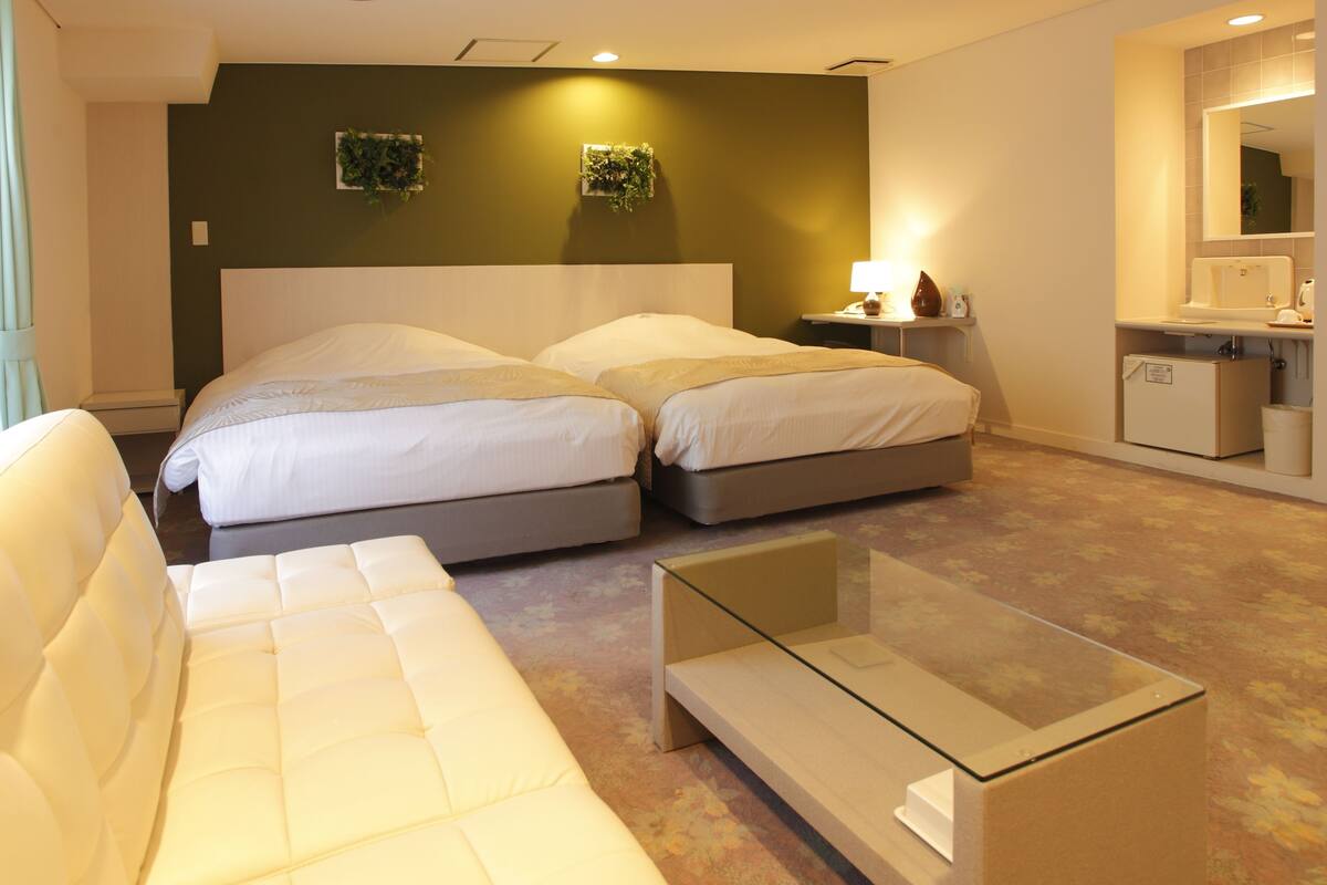 Habitación de hotel con dos camas, una mesita de vidrio, un sofá, una mesita de noche con lámpara y un área pequeña de baño.