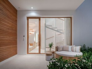 A modern interior with a wooden floor, a seating area with cushions, a staircase, and a glass door leading to a bathroom.