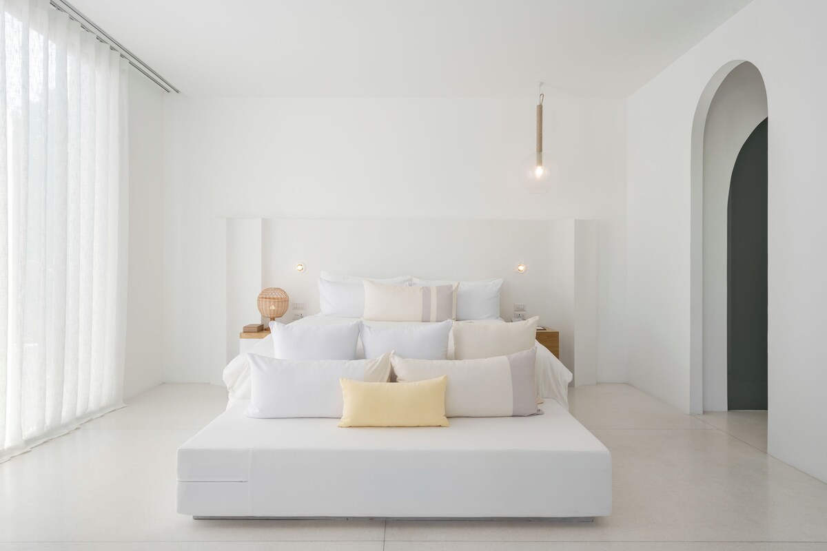 A modern, minimalist bedroom with a large bed, white bedding, and a simple nightstand with a lamp.