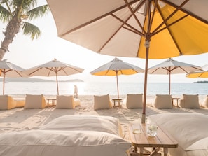 A beachside lounge with multiple umbrellas and lounge chairs, a wooden table, and a view of the sea. - SALA Samui Chaweng Beach Resort (Koh Samui)