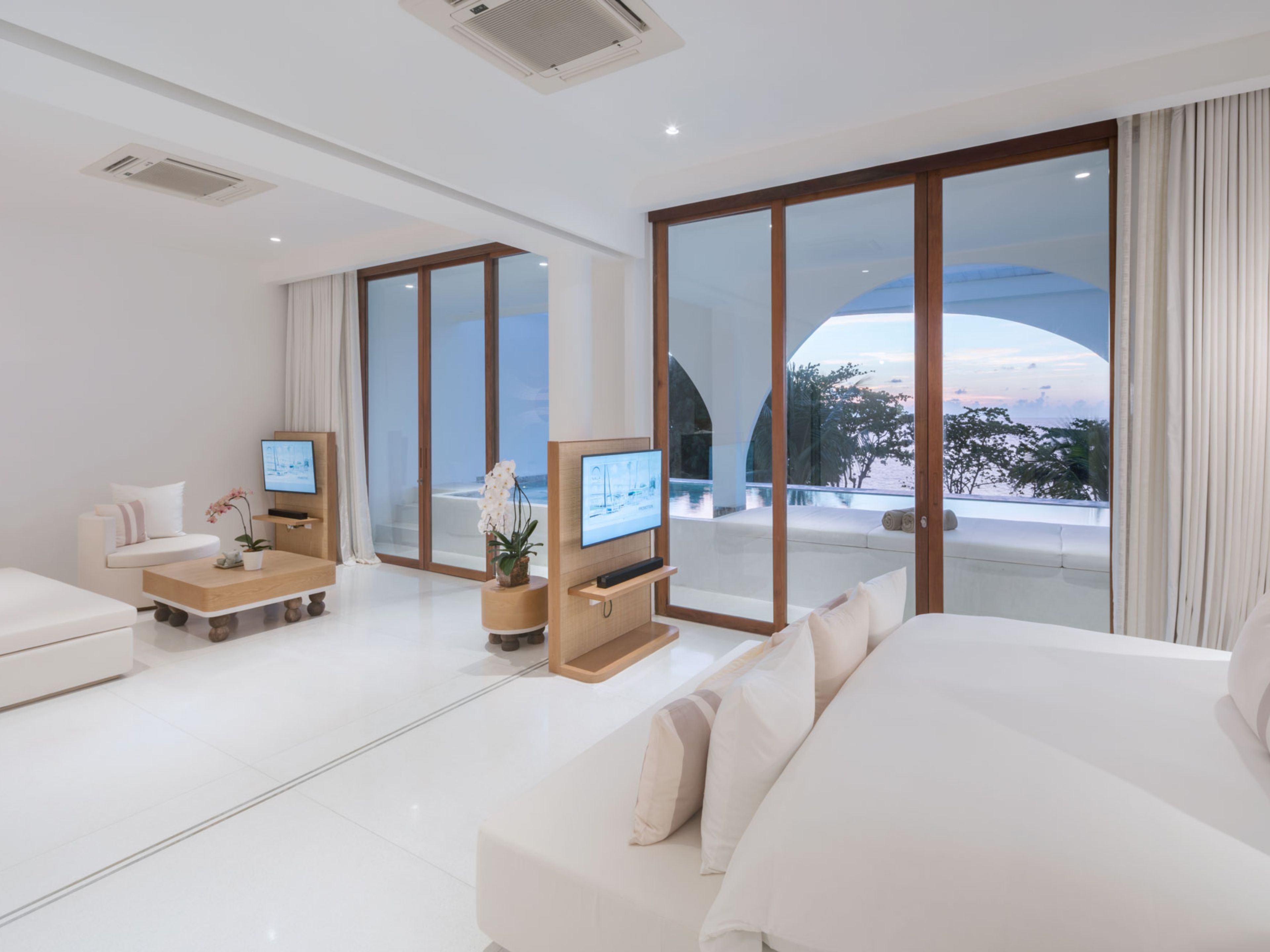 A modern living room with a large sofa, a coffee table, a TV, and a view of a pool and trees.
