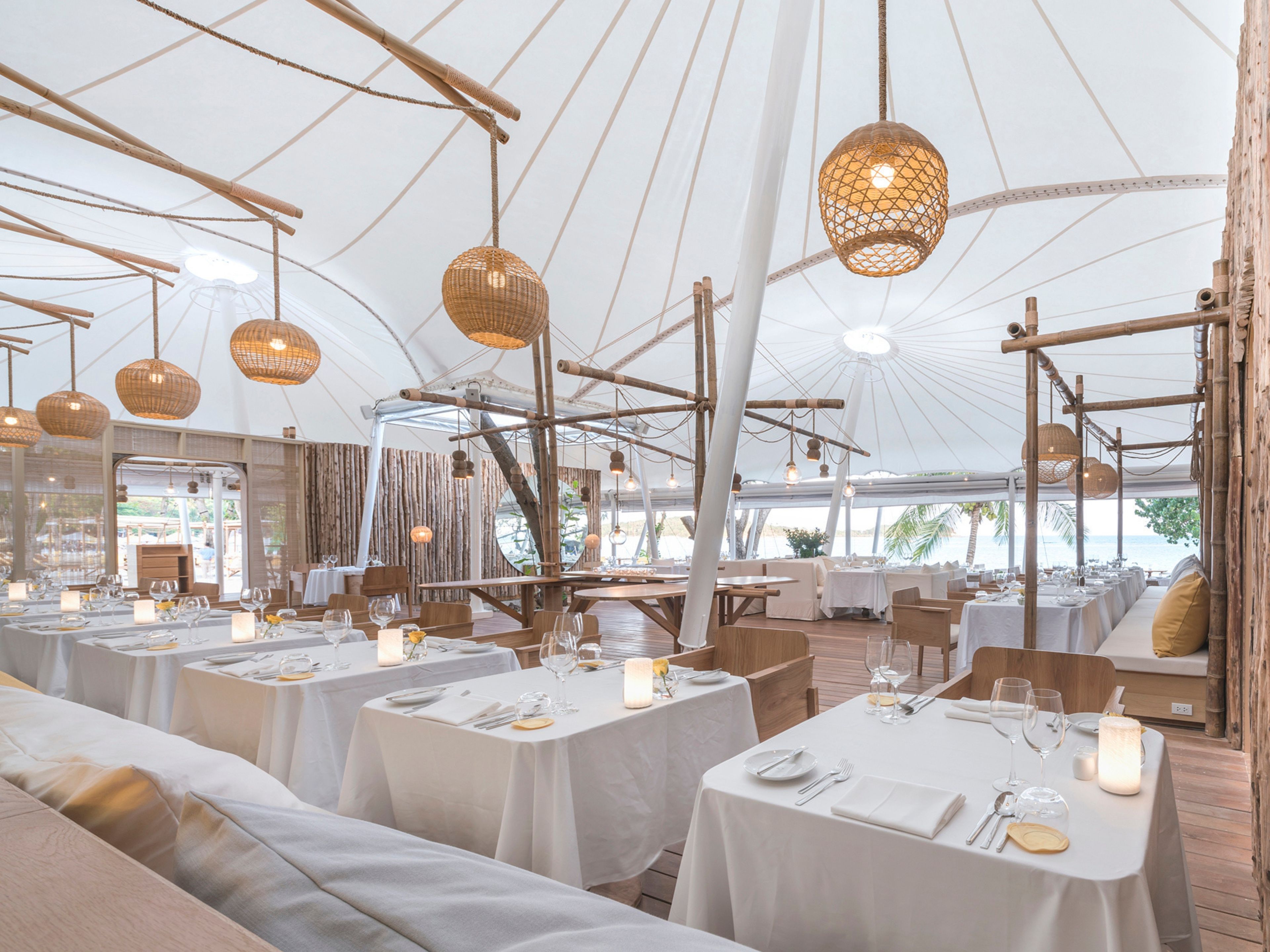 Un área de comedor con carpas, mesas redondas, manteles blancos y lámparas de mimbre colgantes.