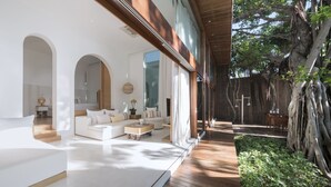 A modern interior with a large white sofa, a coffee table, and a dining area with a wooden table and chairs. There is a large window providing natural light and a view of the outdoors. - SALA Samui Chaweng Beach Resort (Koh Samui)