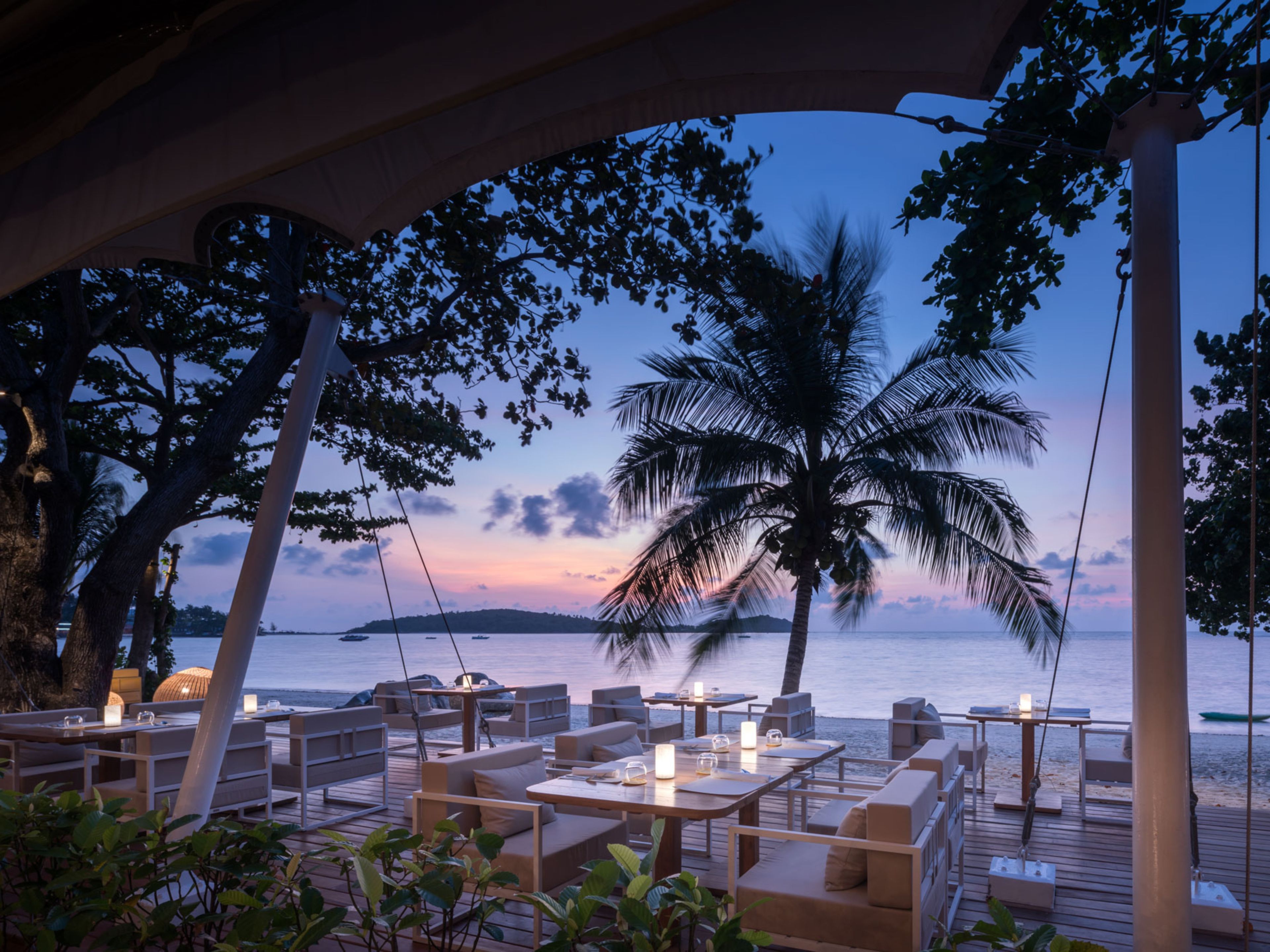Un área de comedor al aire libre con mesas y sillas, con vista a una playa con palmeras y un atardecer.
