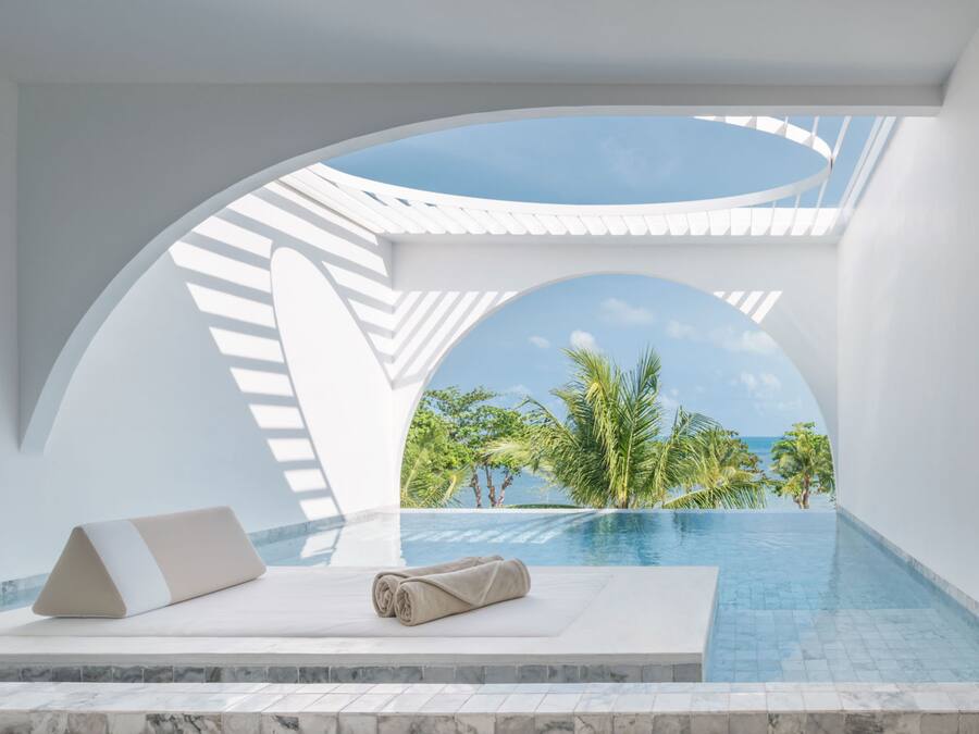 Un área de piscina moderna con vistas despejadas al océano y palmeras, que incluye una estructura en arco blanca y un espacio lounge con toalla y almohada.