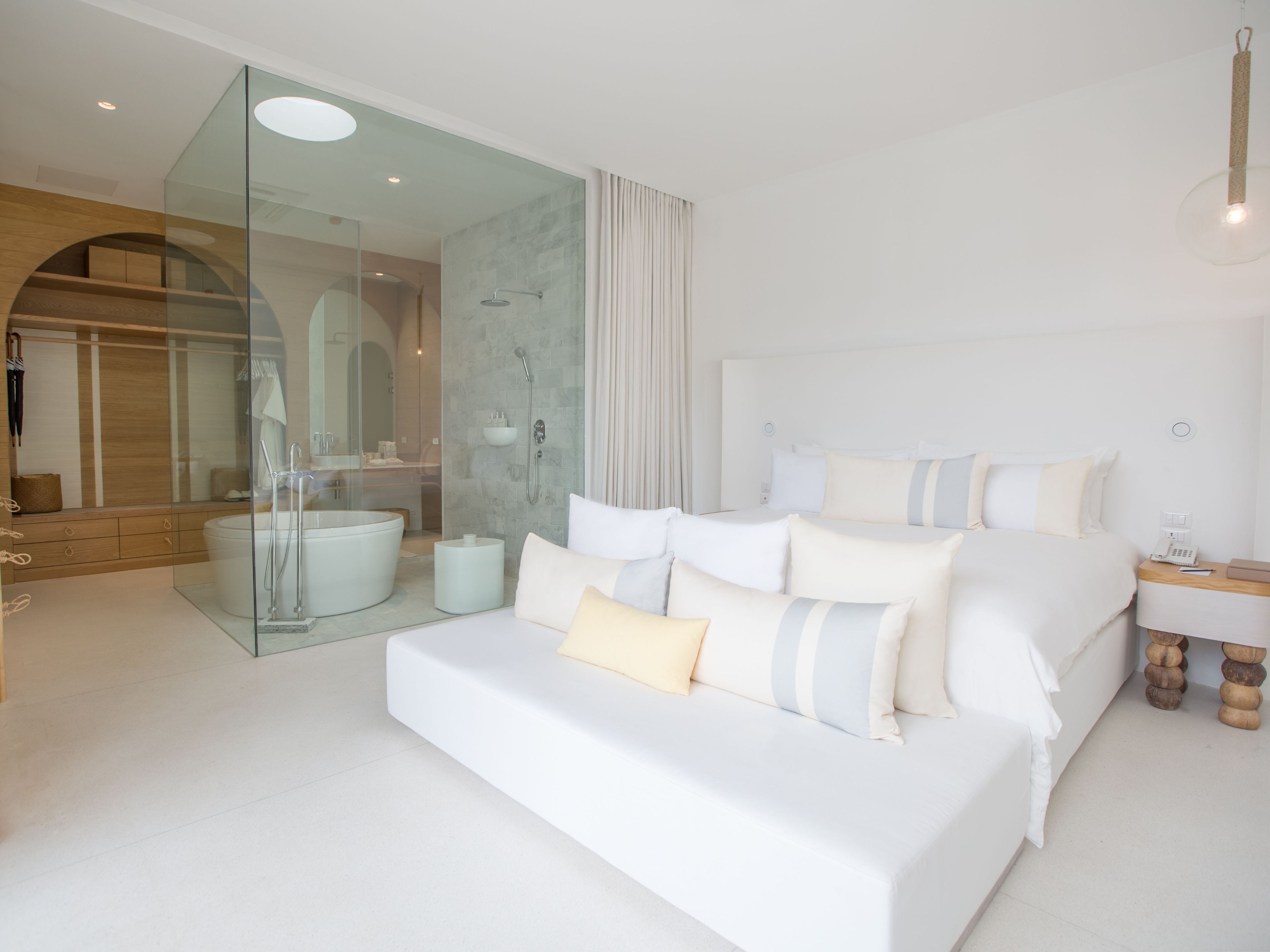 Ein modernes Hotelzimmer mit einer Glaskabine zur Dusche, einem großen Bett mit vielen Kissen und einem Nachttisch mit Lampe und Telefon.