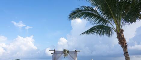 A beach wedding setup with a canopy, floral decorations, and white chairs.