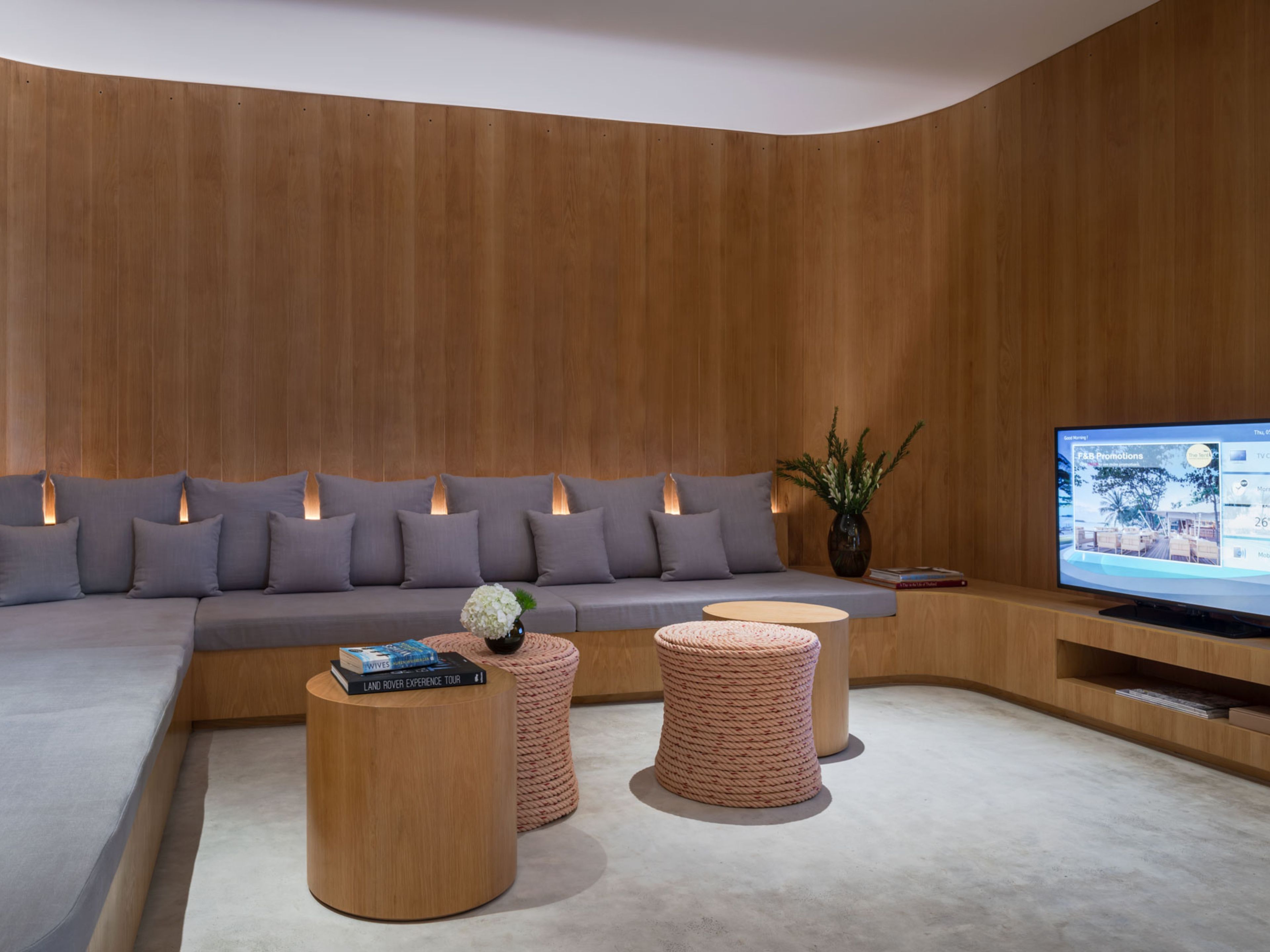A modern living room with a curved wooden wall, a grey sofa with multiple cushions, a flat-screen TV, and two round wooden tables with books and a vase.