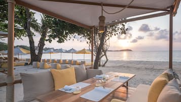A beachside dining area with a wooden table, cushioned seating, and umbrellas.
