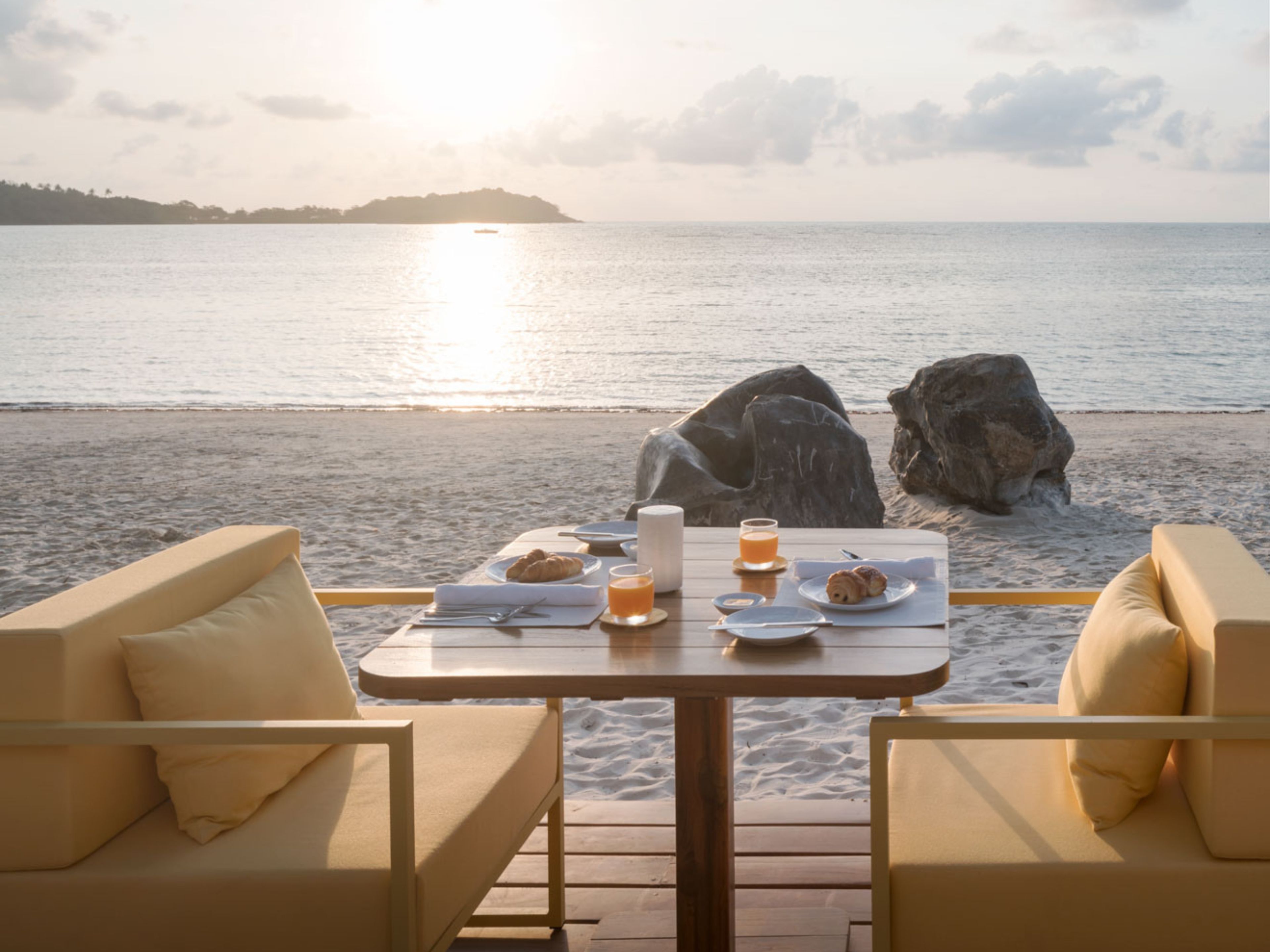 A beachside table with breakfast items, two chairs, and a view of the sea.