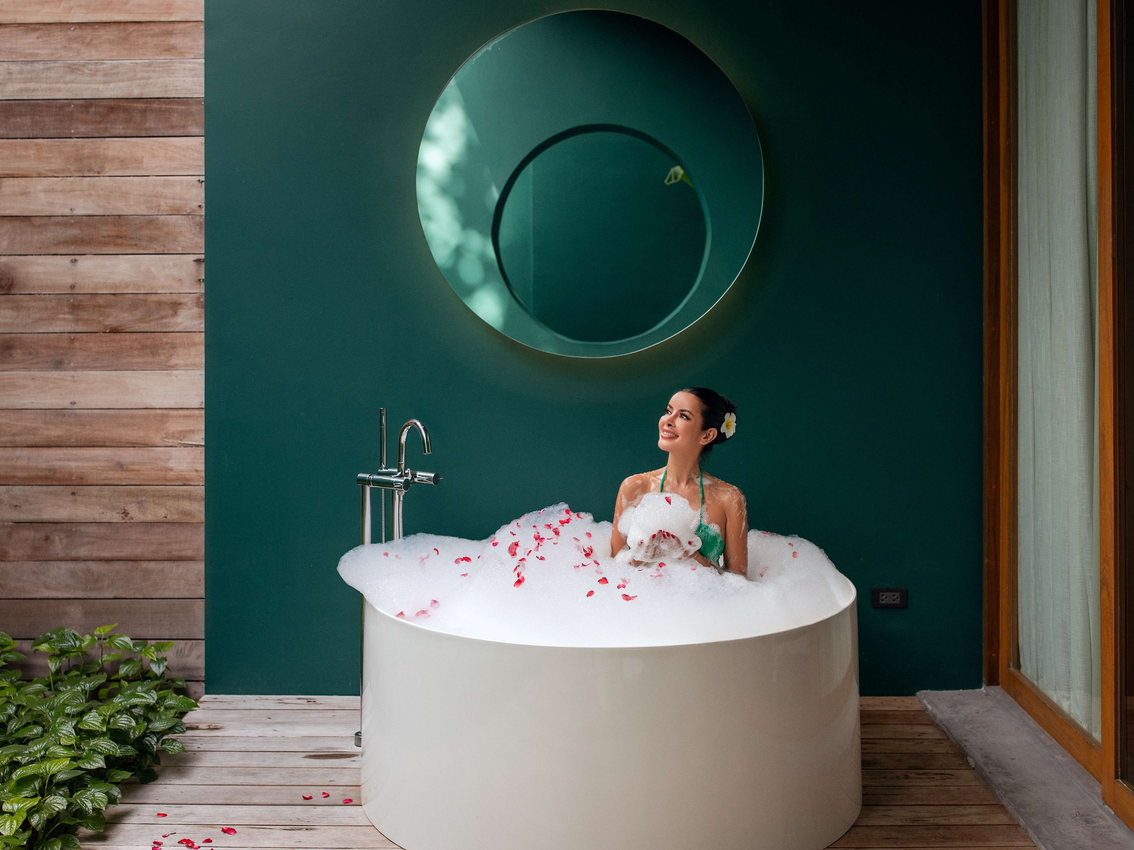 A person in a bathtub filled with bubbles and rose petals, with a wooden floor and a green wall with a circular mirror.