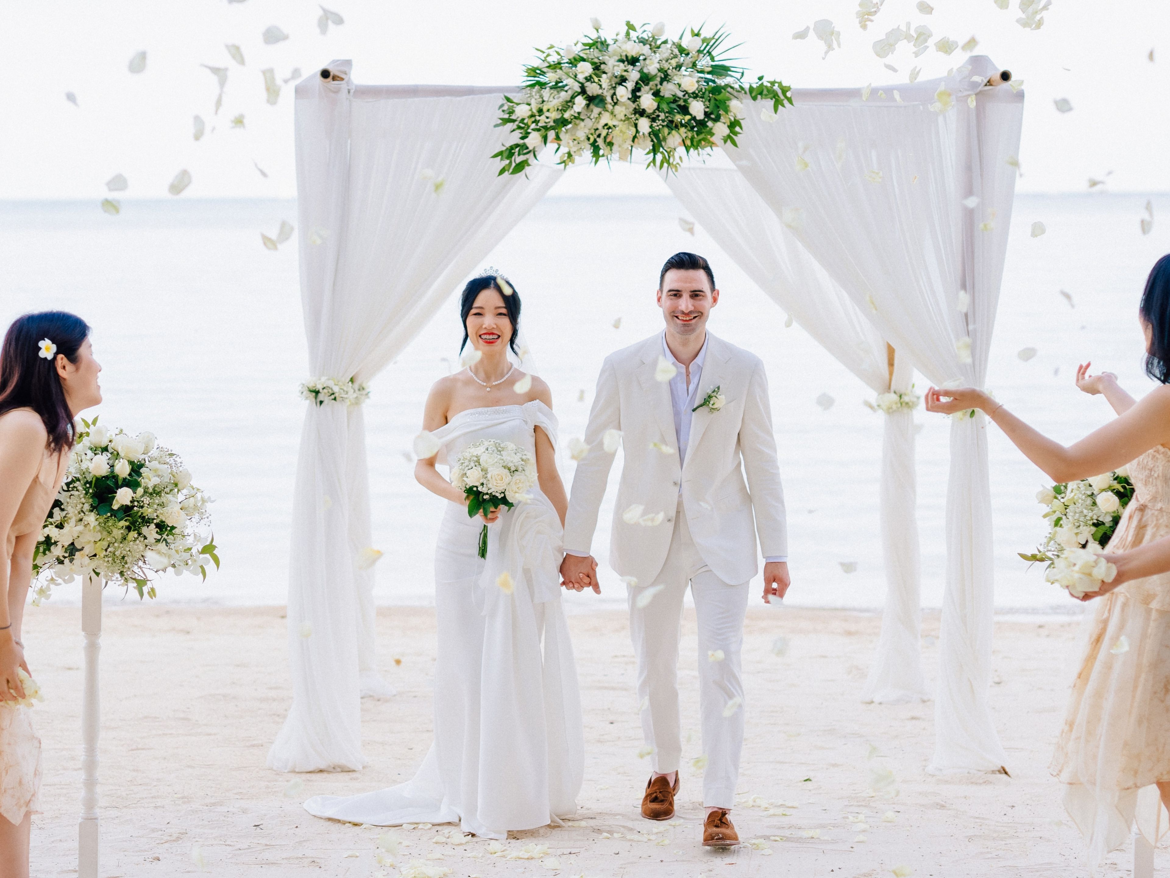 Ceremonia de boda en la playa con un arco blanco e invitados sosteniendo ramos de flores.