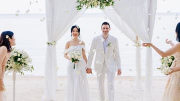 Ceremonia de boda en la playa con un arco blanco e invitados sosteniendo ramos de flores.