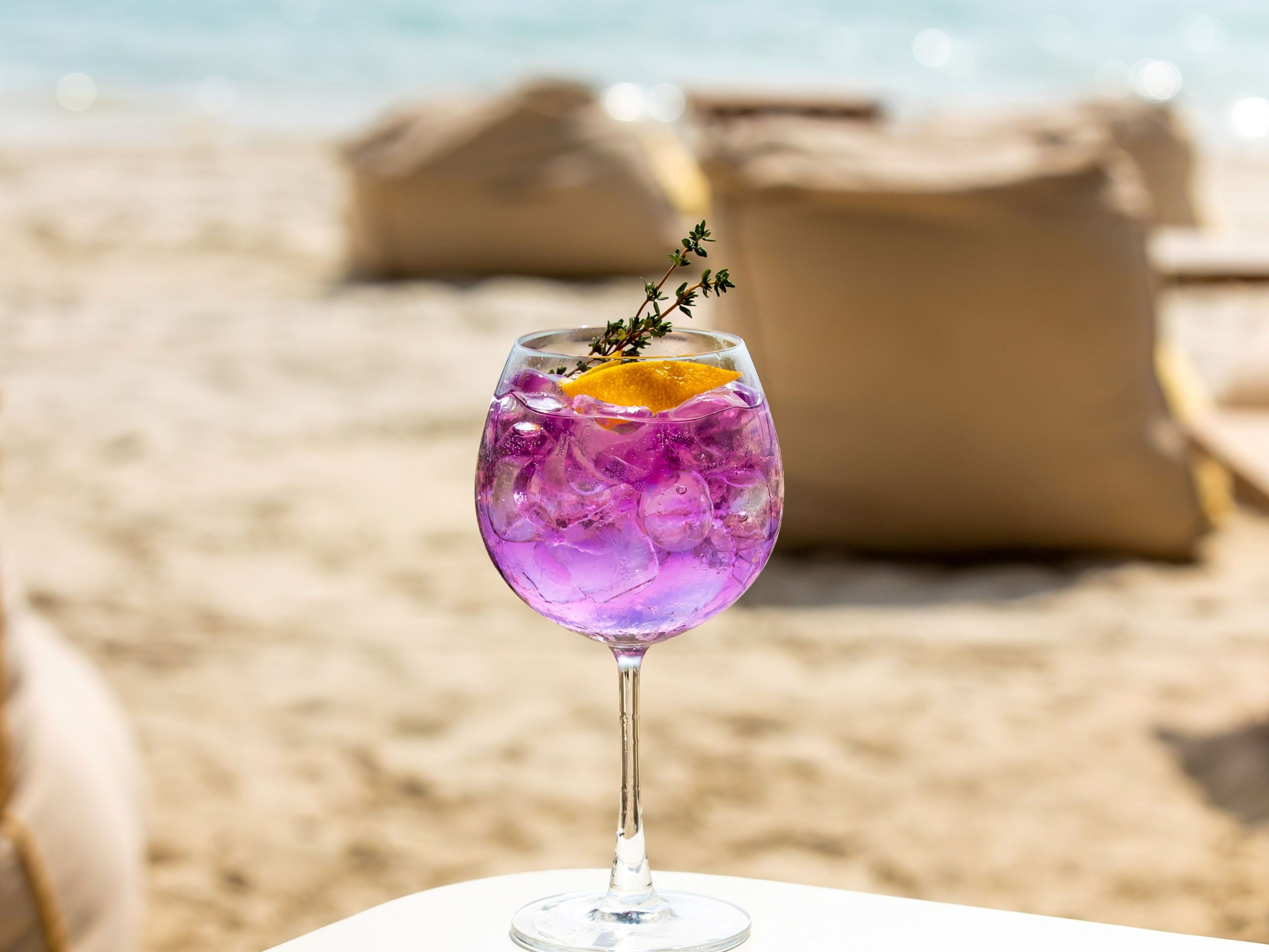 Un vaso de bebida morada con una rodaja de naranja y una ramita de tomillo sobre una mesa blanca con el mar de fondo.