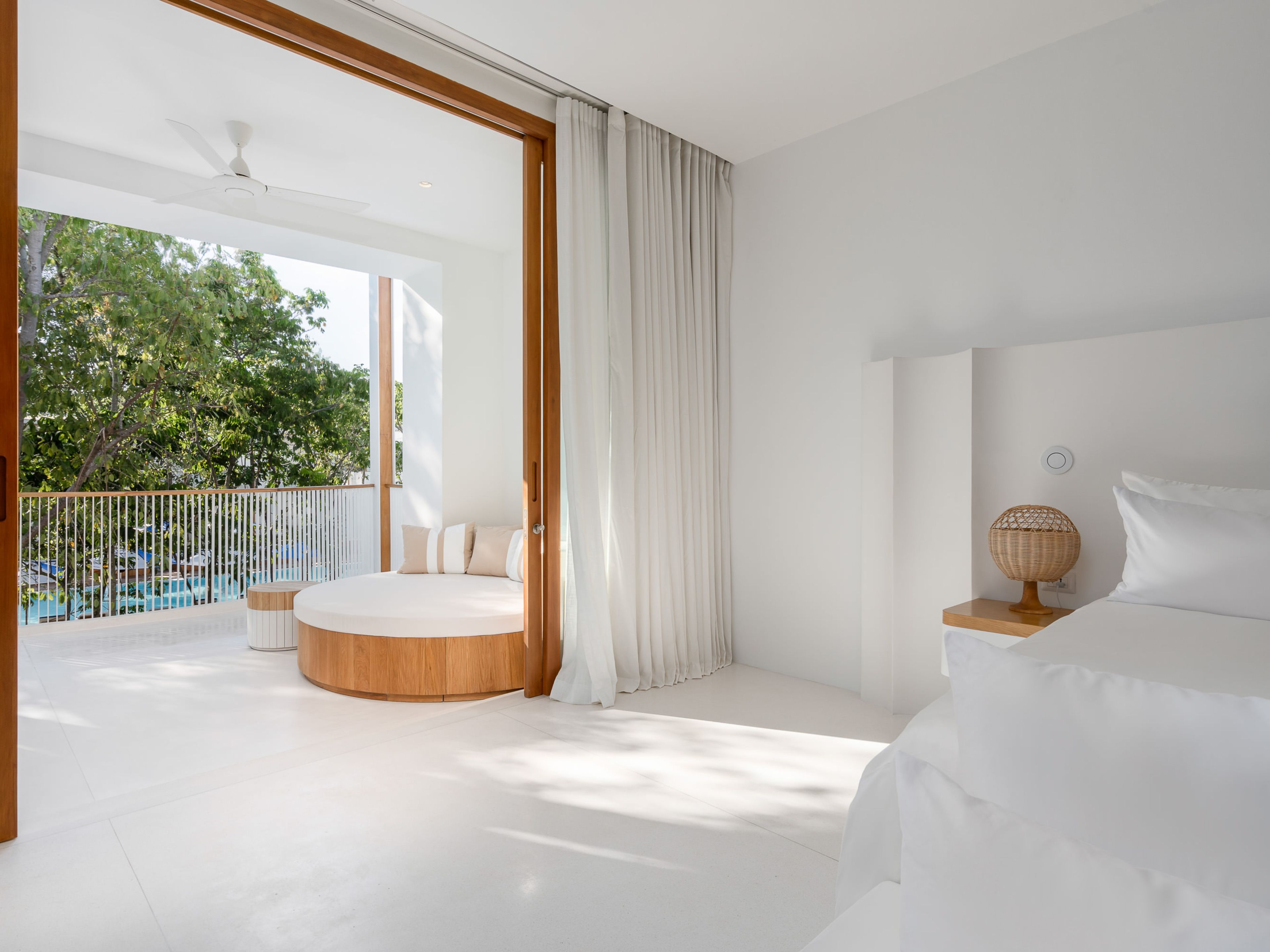 A modern bedroom with a circular wooden bed, a bedside table with a woven lamp, and a view of a pool and trees.