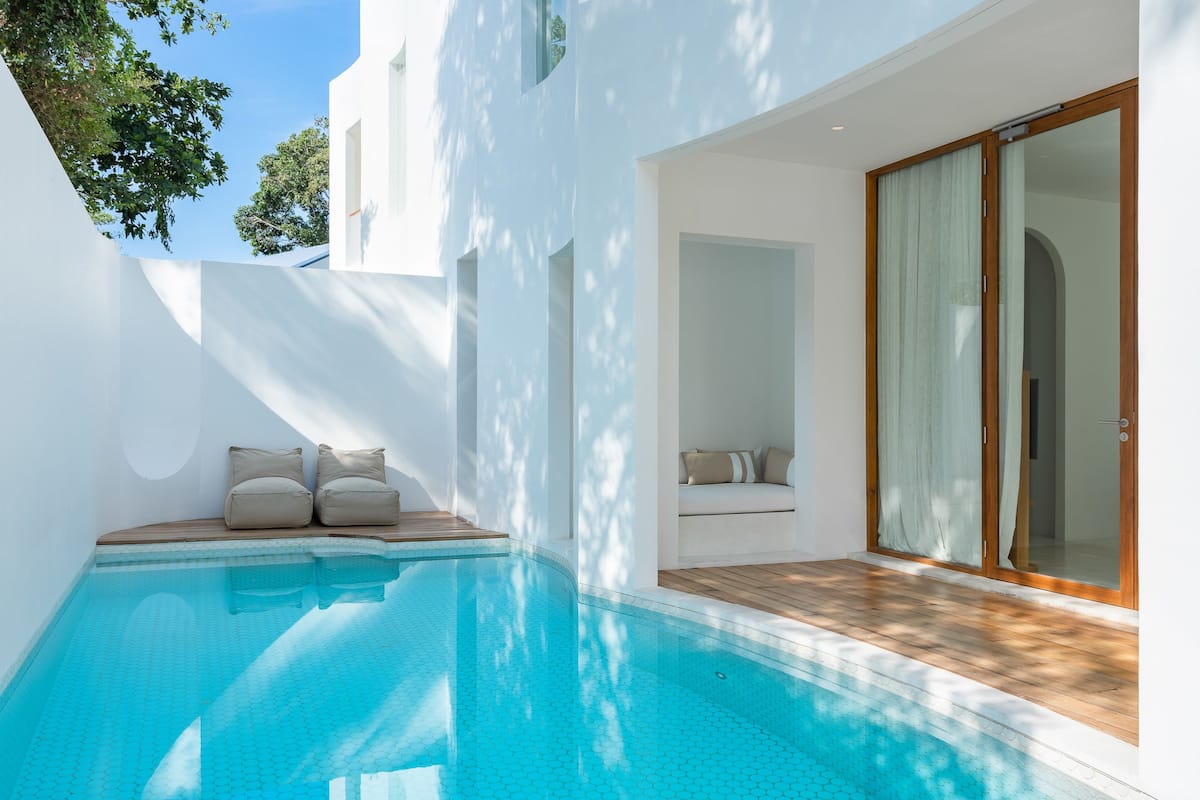 Un espace piscine moderne avec une piscine à l’eau d’un bleu limpide, une terrasse en bois et un bâtiment blanc doté de grandes fenêtres et de portes-fenêtres coulissantes.