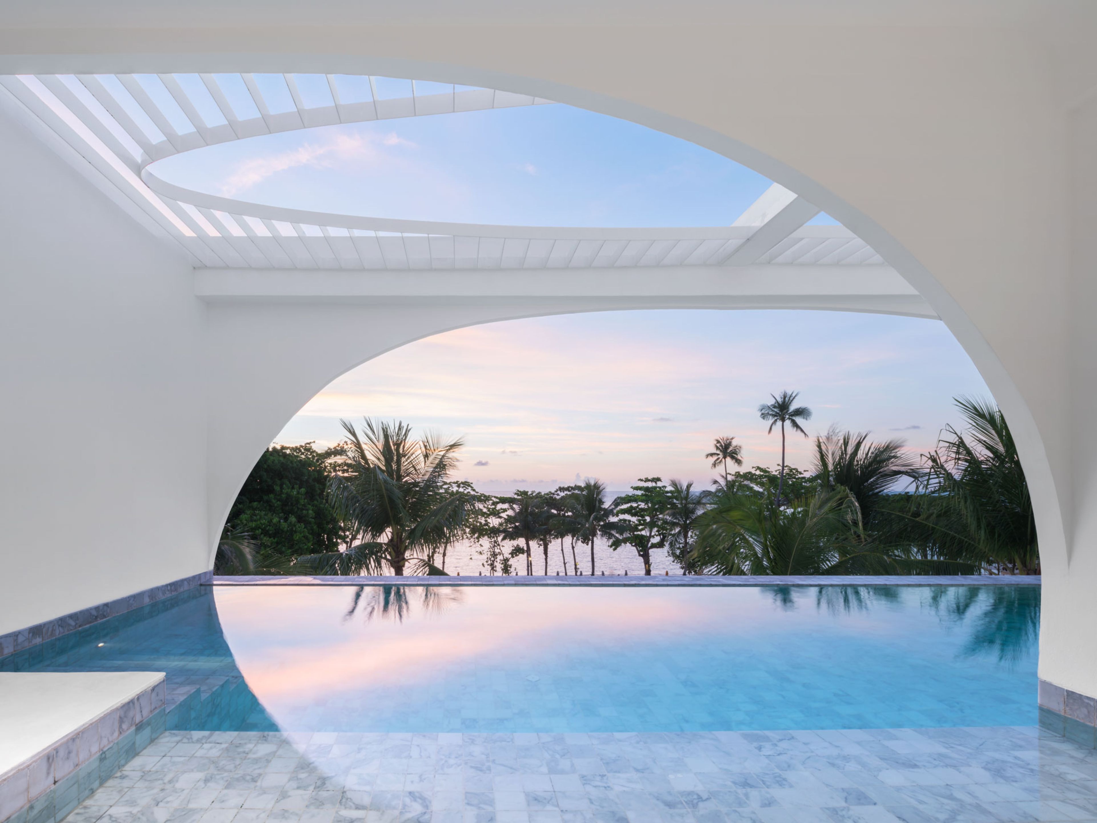 Vista despejada de una piscina con una estructura blanca curva en la parte superior, que refleja el cielo y palmeras circundantes.