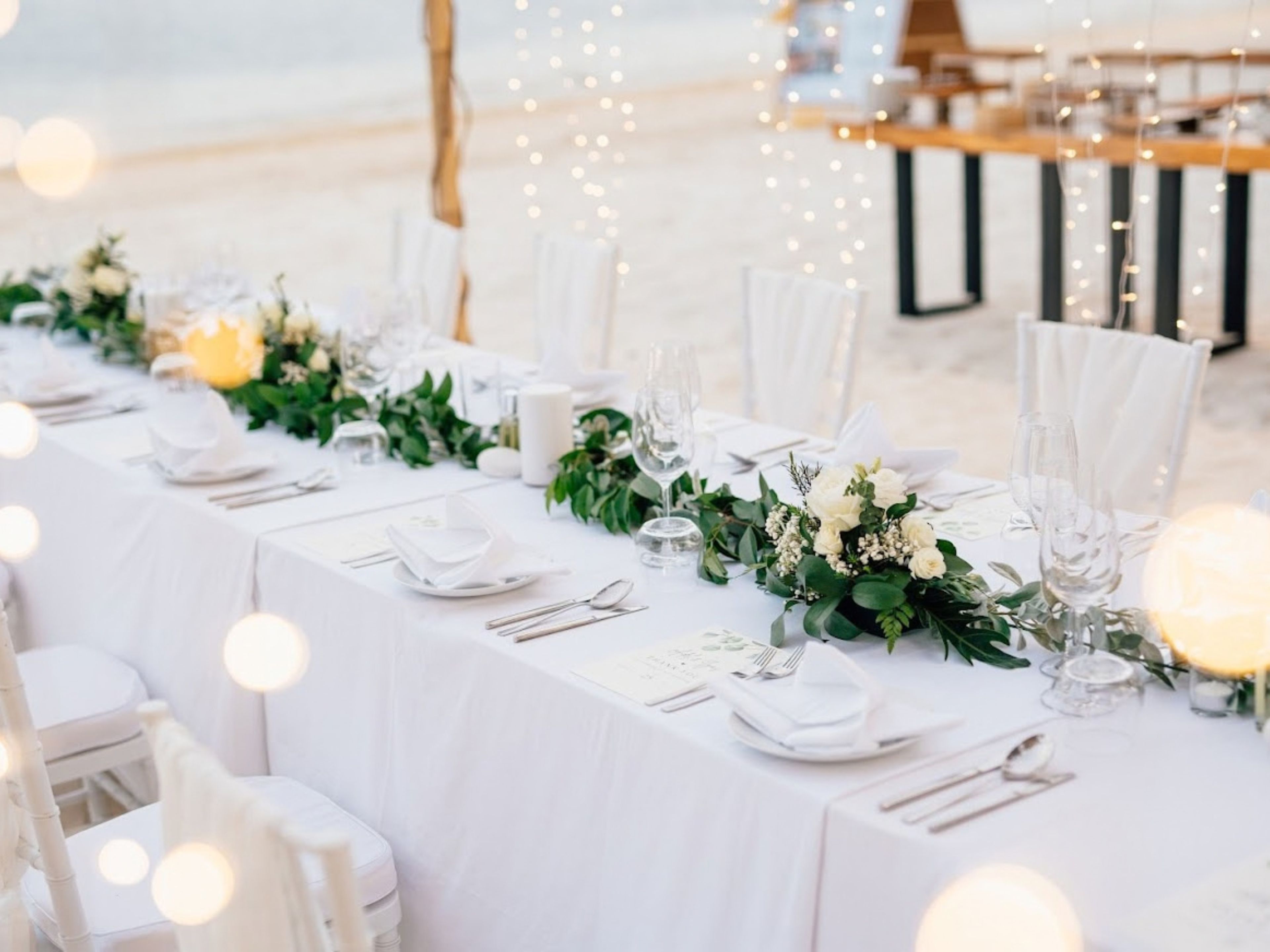 A beachside dining setup with white tablecloths, neatly arranged cutlery, and floral centerpieces.