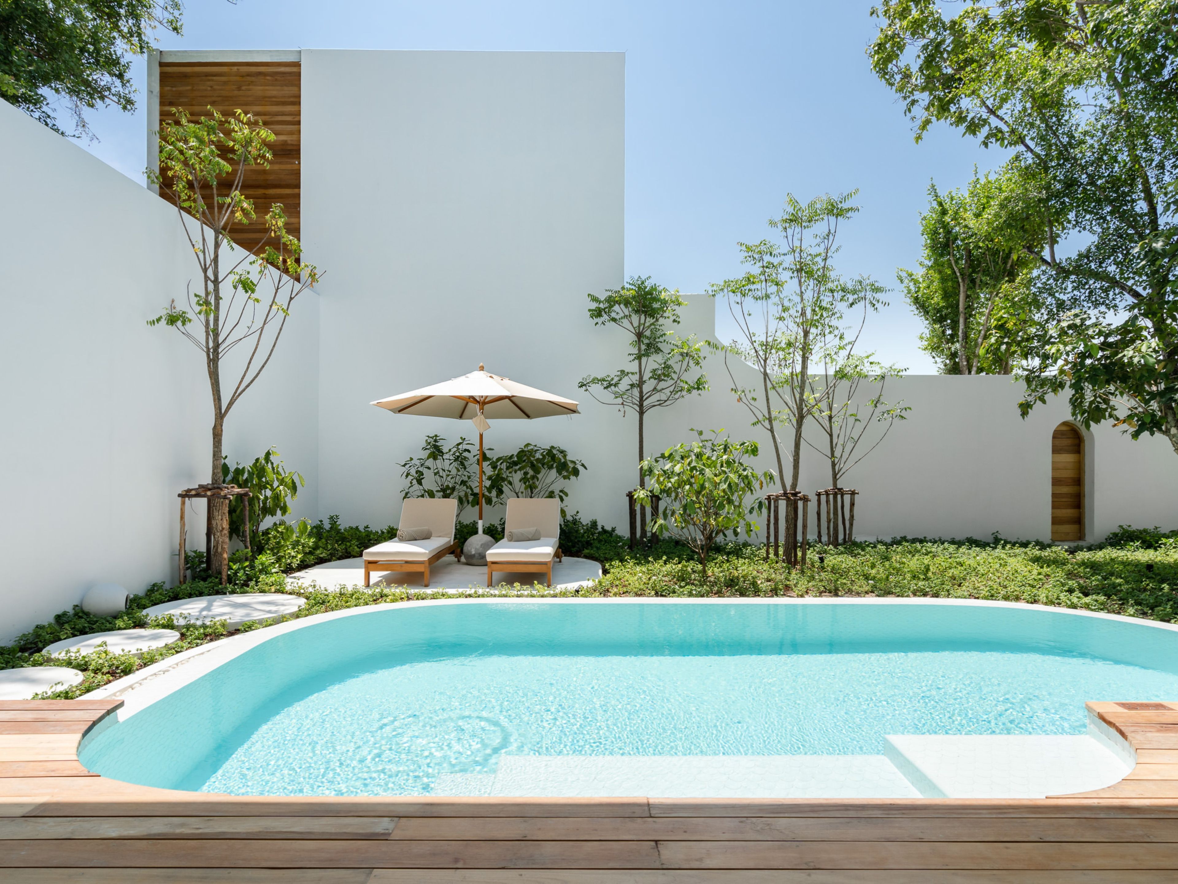 A swimming pool with two lounge chairs and an umbrella, surrounded by greenery and a white wall.