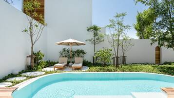 A swimming pool with two lounge chairs and an umbrella, surrounded by greenery and a white wall.