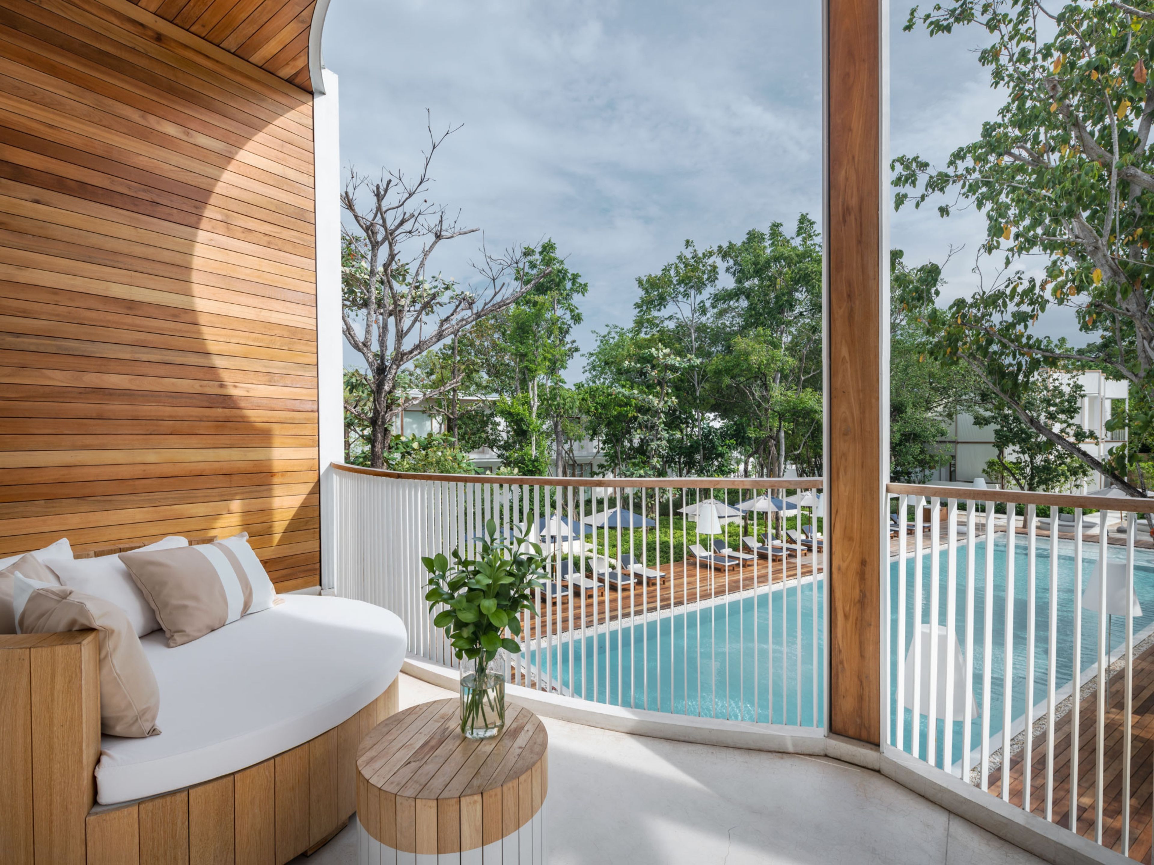 A balcony with a wooden floor, a white cushioned seating area, a small round table with a vase, and a view of a swimming pool and greenery.