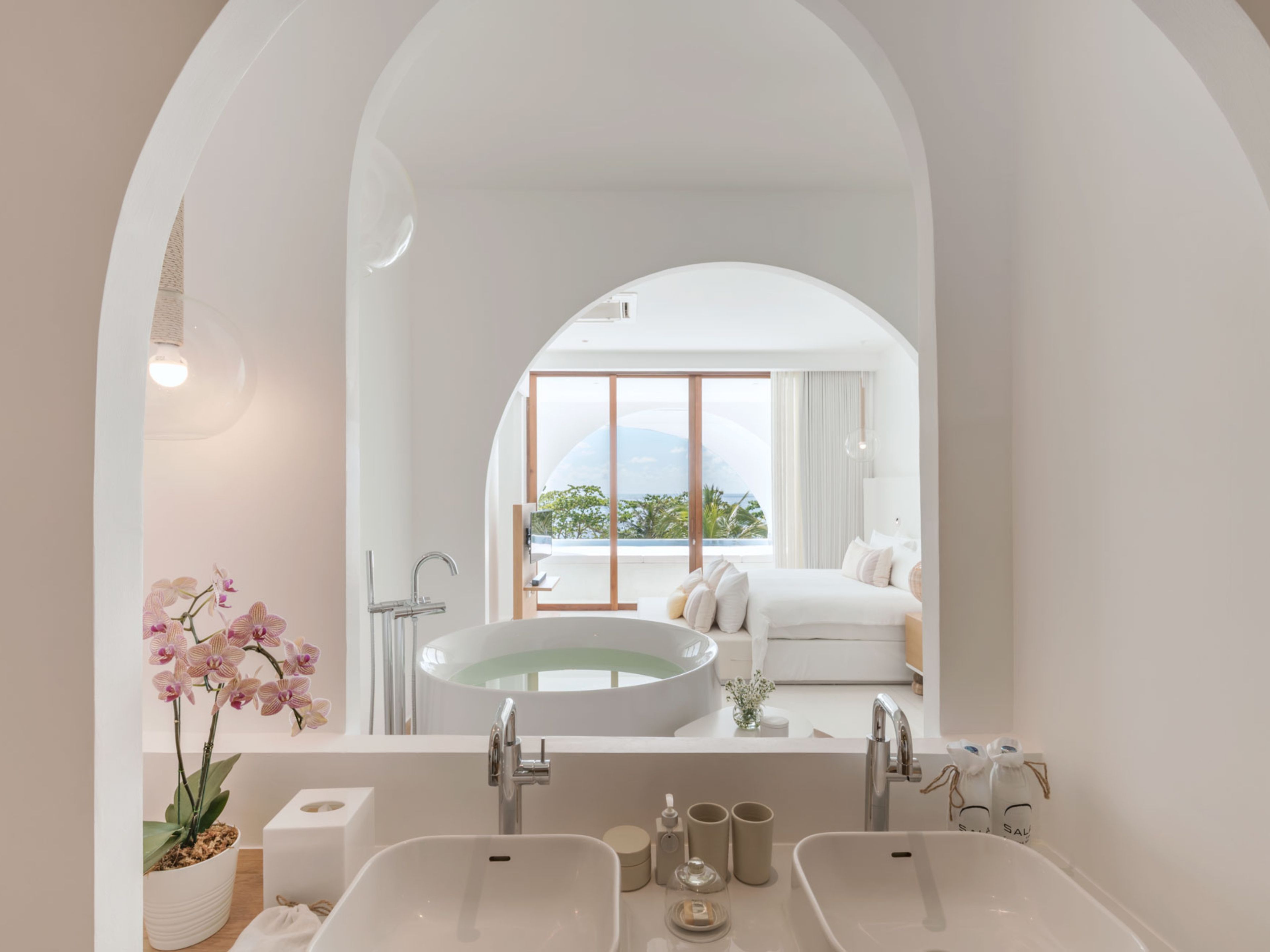 A modern bathroom with a large oval sink, a mirror, and a view of a bedroom with a bed and window.