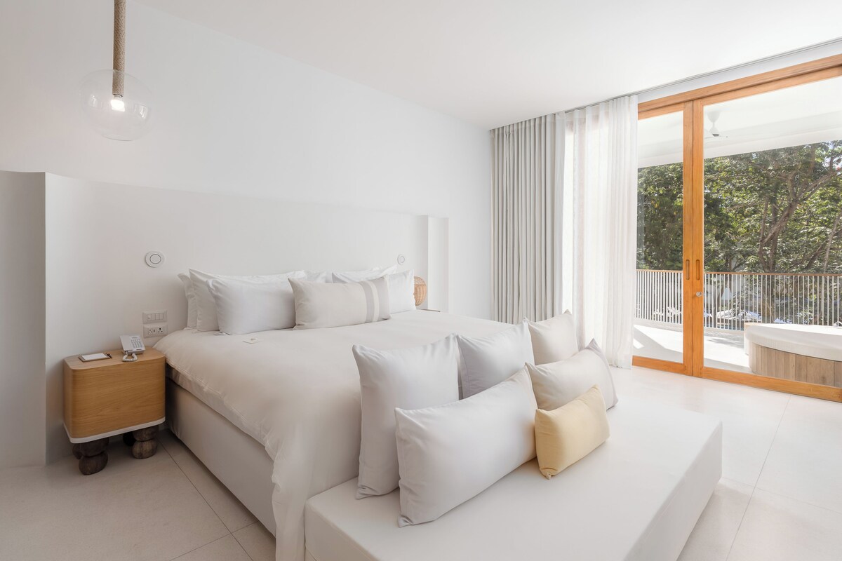 A modern bedroom with a large bed, bedside table, and a view of the outdoors.