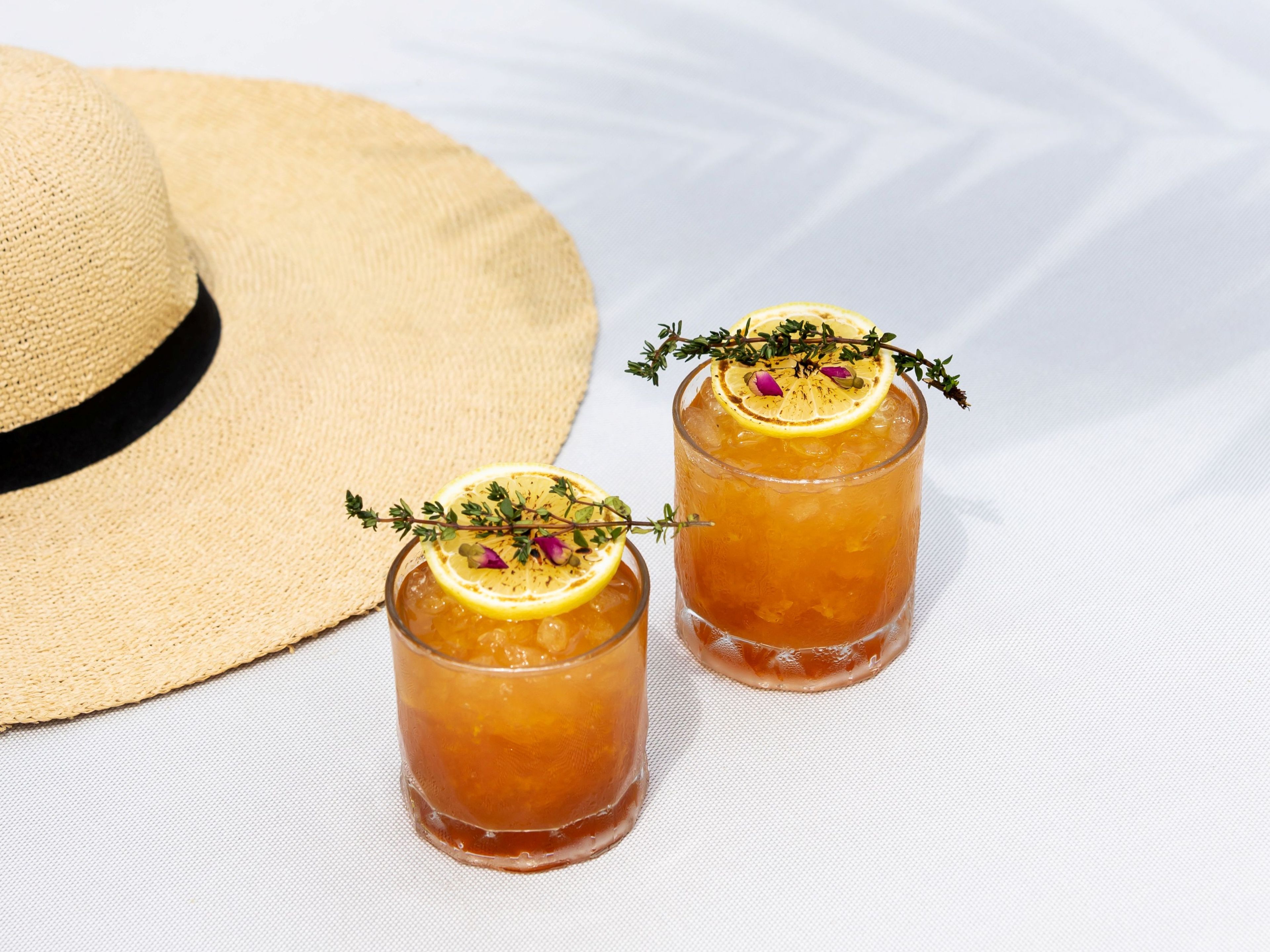 Dos vasos de bebida color ámbar con rodajas de limón y hierbas, y un sombrero de paja.