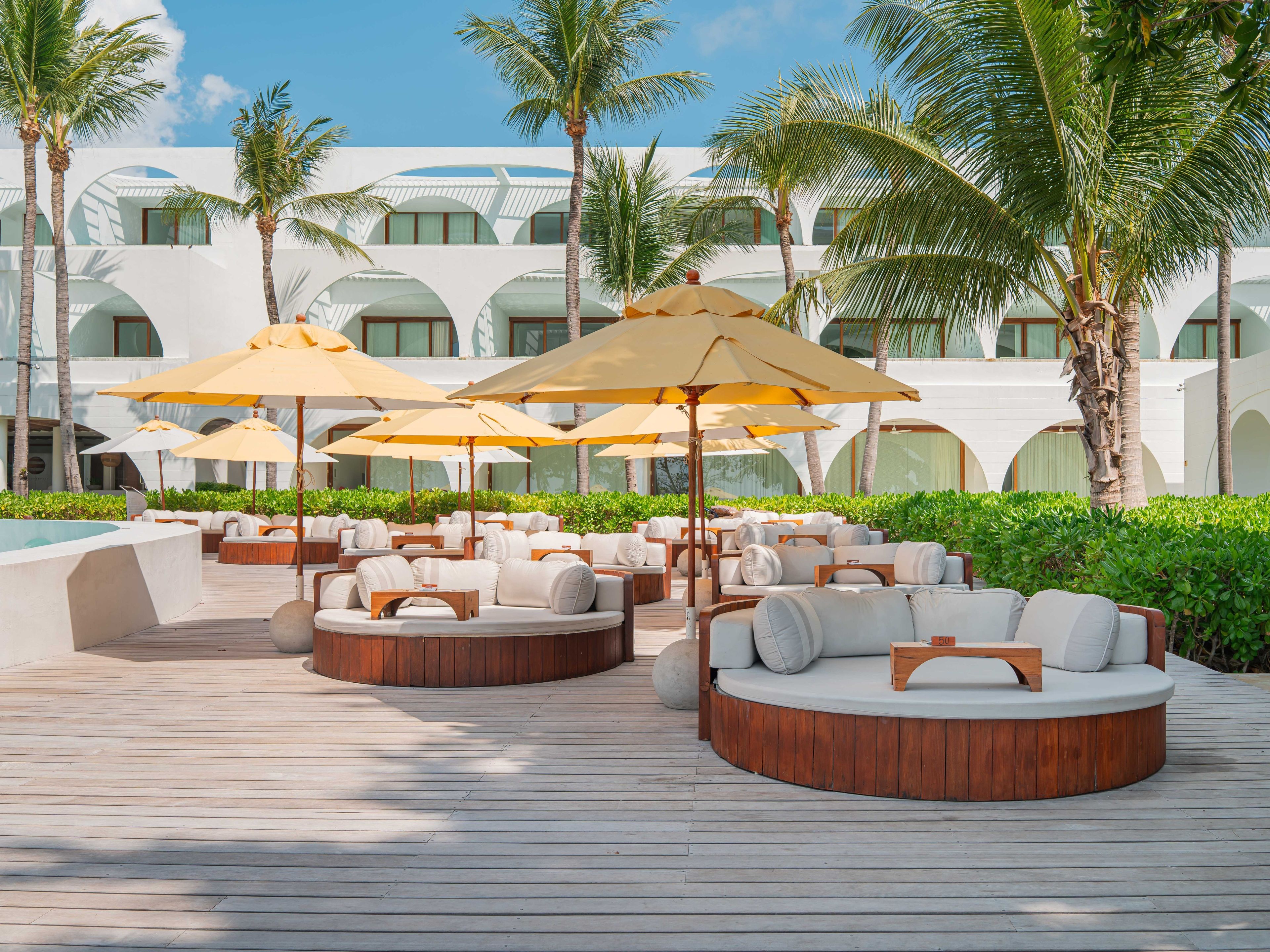 Zona de estar al aire libre en un resort con sofás de madera redondos y sombrillas beige, rodeada de palmeras y un edificio blanco con ventanas arqueadas.