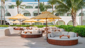 Zona de estar al aire libre en un resort con sofás de madera redondos y sombrillas beige, rodeada de palmeras y un edificio blanco con ventanas arqueadas.