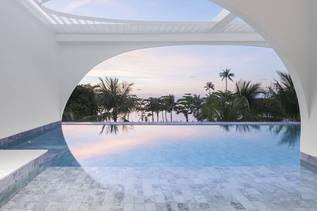 Une piscine à débordement moderne offrant une vue dégagée sur l’océan et les palmiers, le tout mis en valeur par une architecture blanche.
