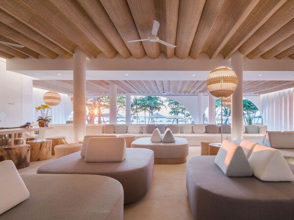 A modern lounge area with a ceiling fan, cushioned seating, and large windows overlooking a beach.