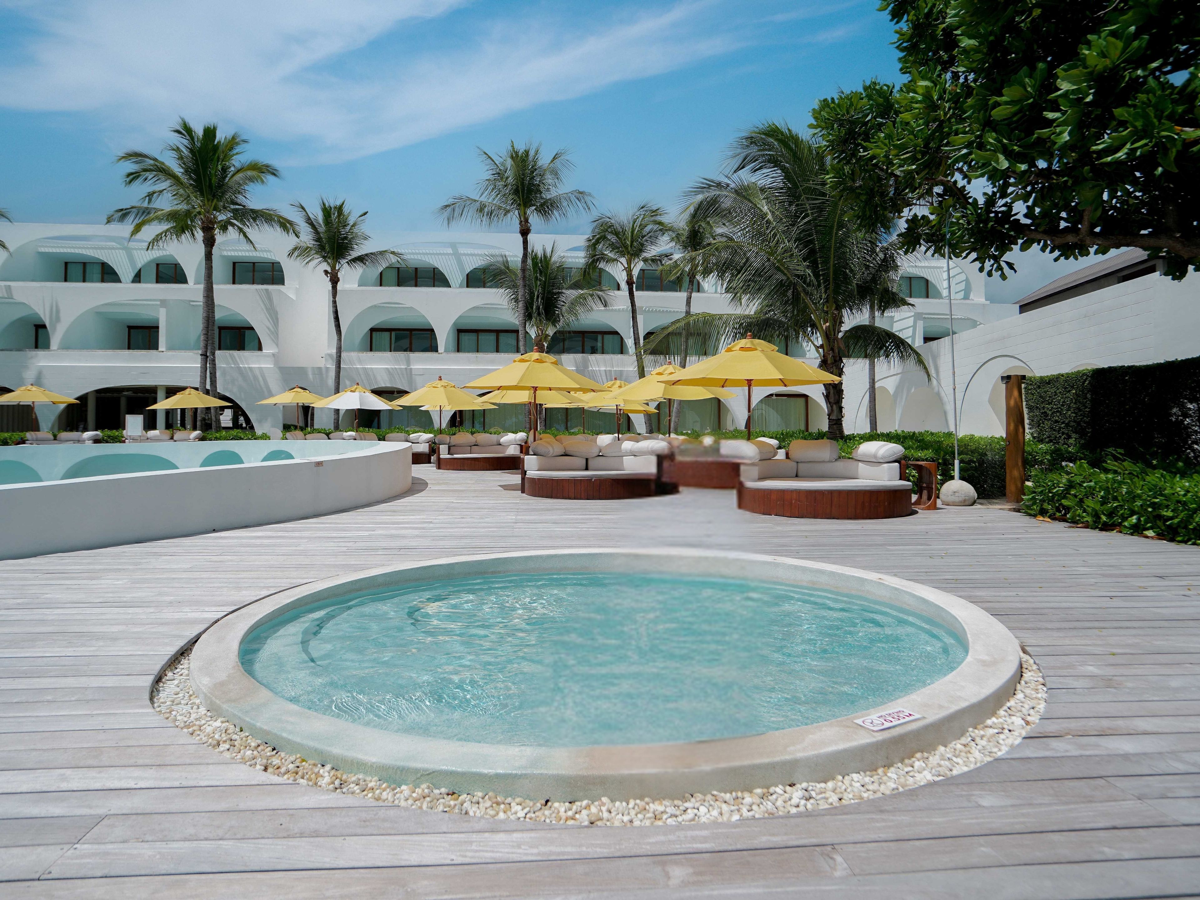 A pool area with lounge chairs and umbrellas, surrounded by palm trees and a building with arched windows.