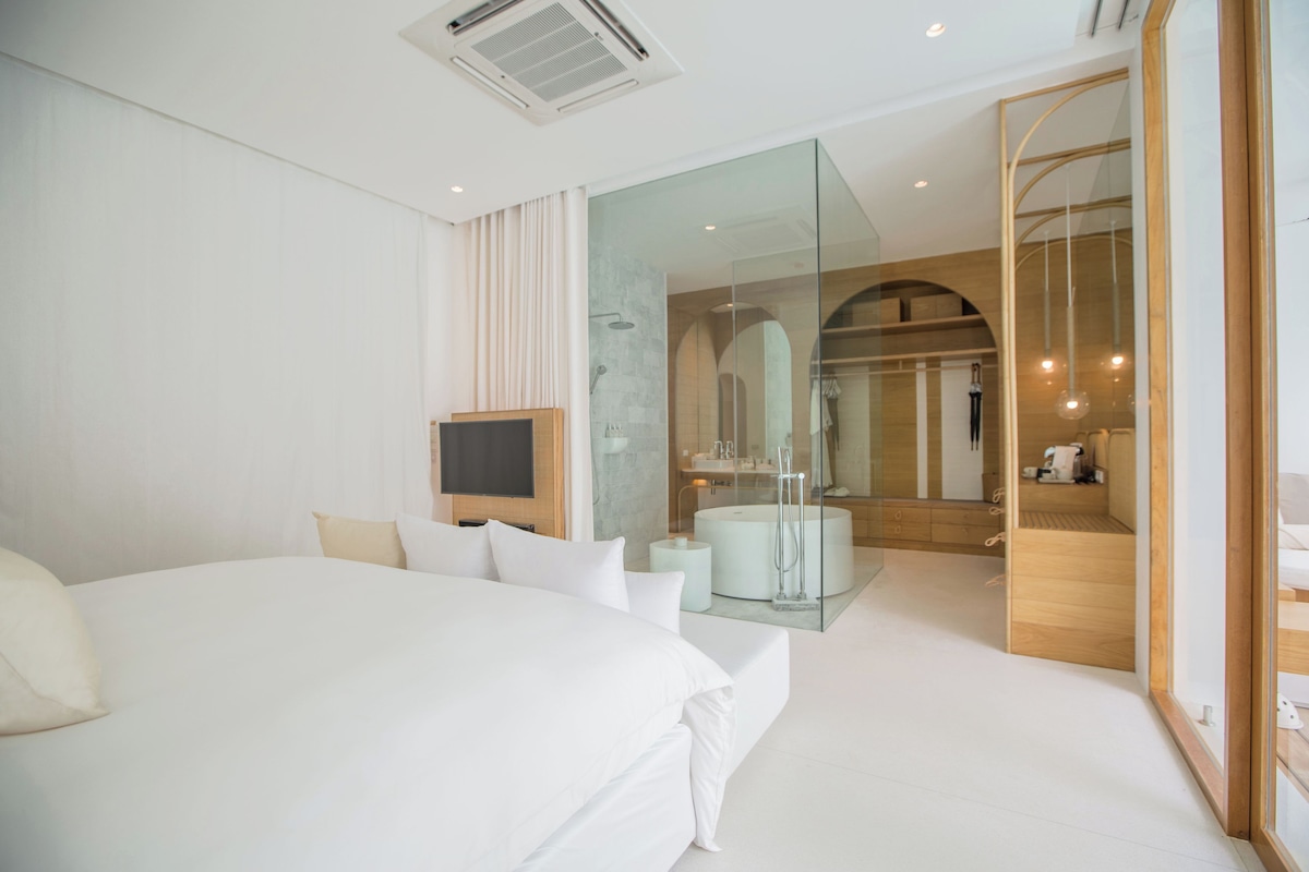 A modern hotel room with a large bed, a television, a glass-enclosed bathroom, and a wooden wardrobe.