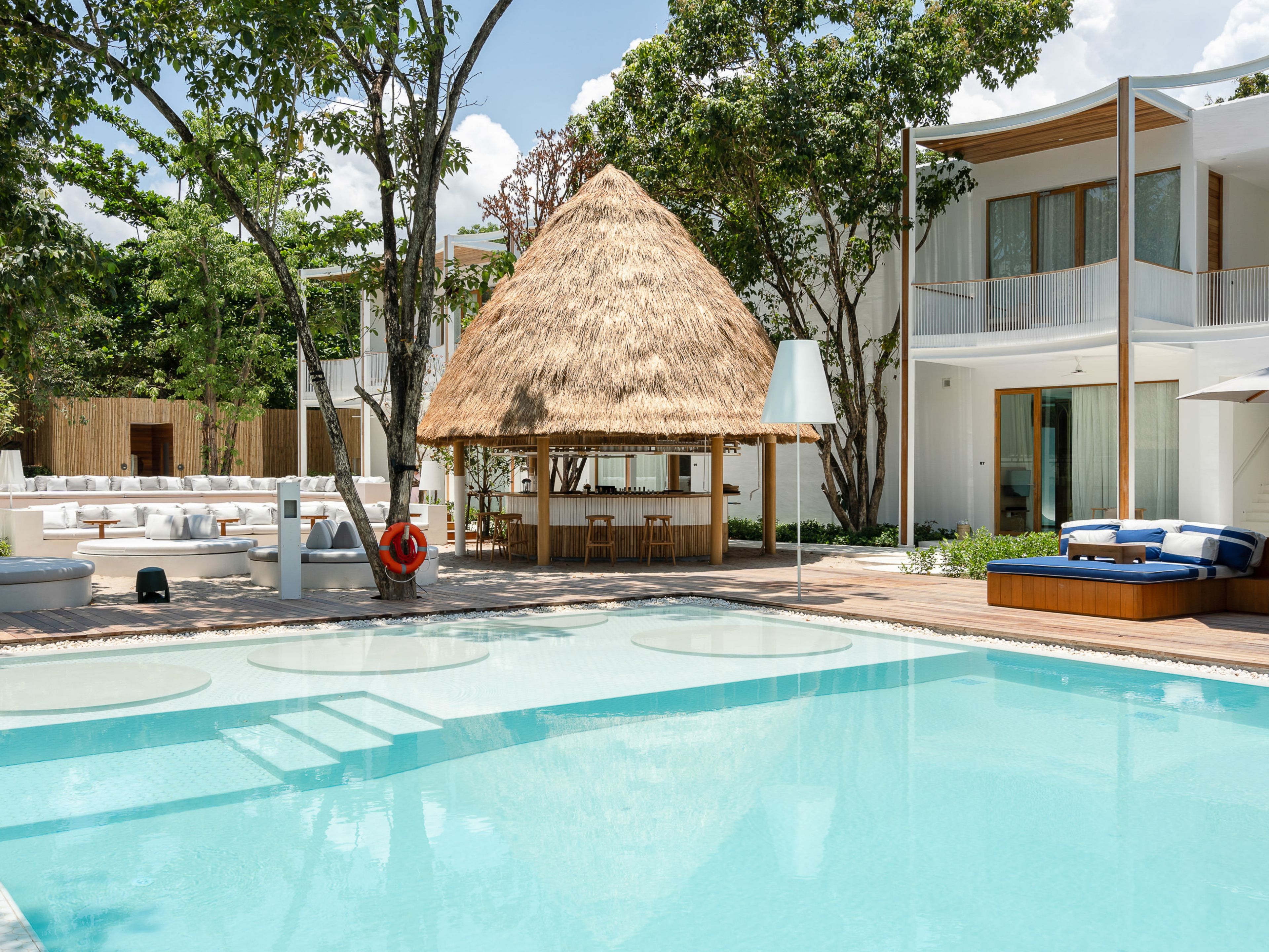 Un área de piscina con una estructura de techo de paja, asientos de descanso y un edificio moderno con grandes ventanales.