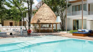 Un área de piscina con una estructura de techo de paja, asientos de descanso y un edificio moderno con grandes ventanales.