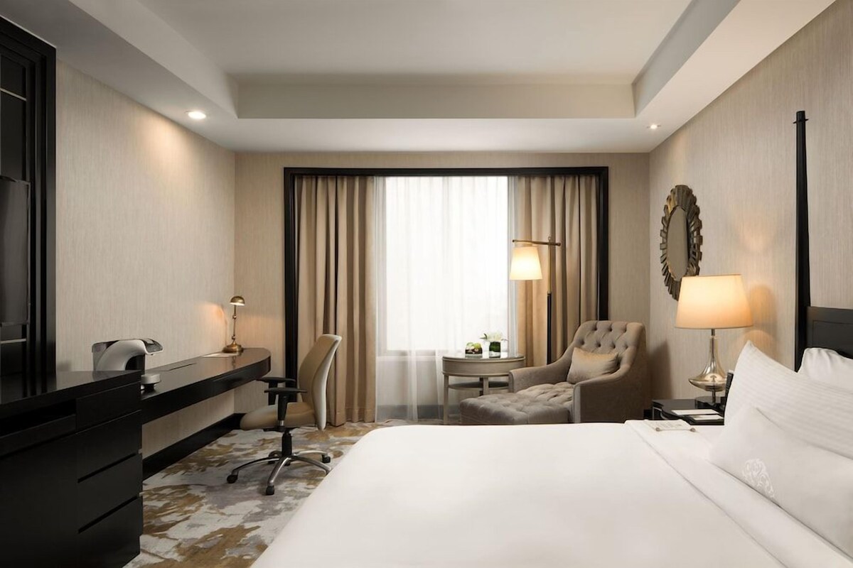 Ein Hotelzimmer mit Bett, Schreibtisch, Stuhl, Lampe und einem großen Fenster mit Vorhängen.