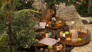 A circular wooden structure with seating and a central umbrella, surrounded by lush greenery and stone flooring.