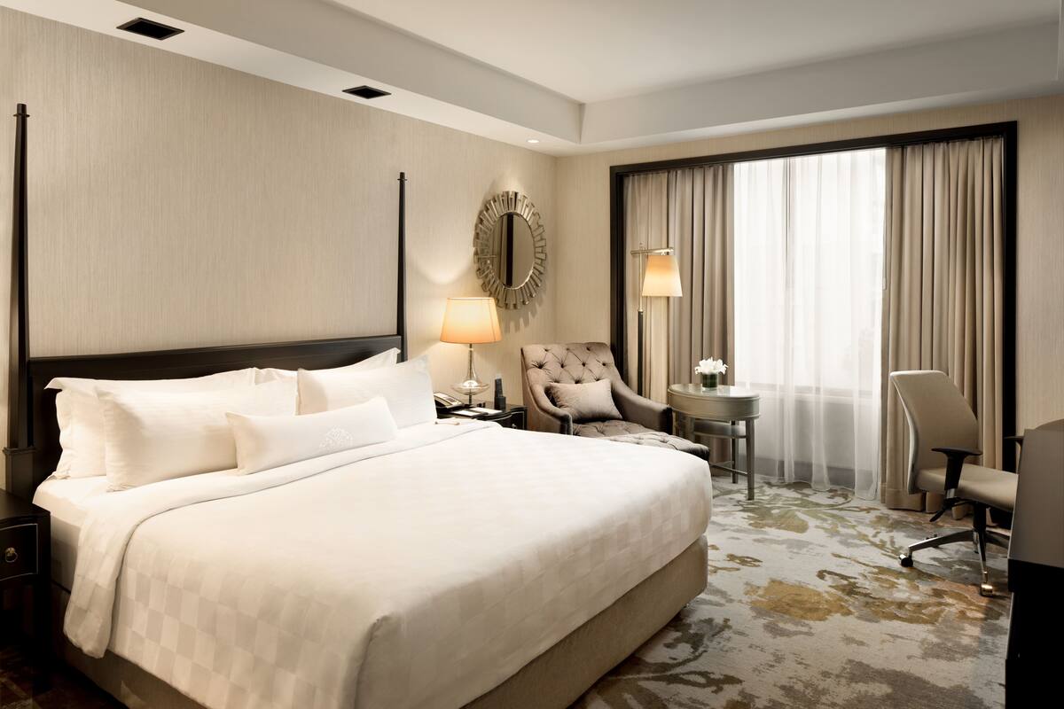 A hotel room with a large bed, a round mirror, two chairs, a small table, and a large window with curtains.