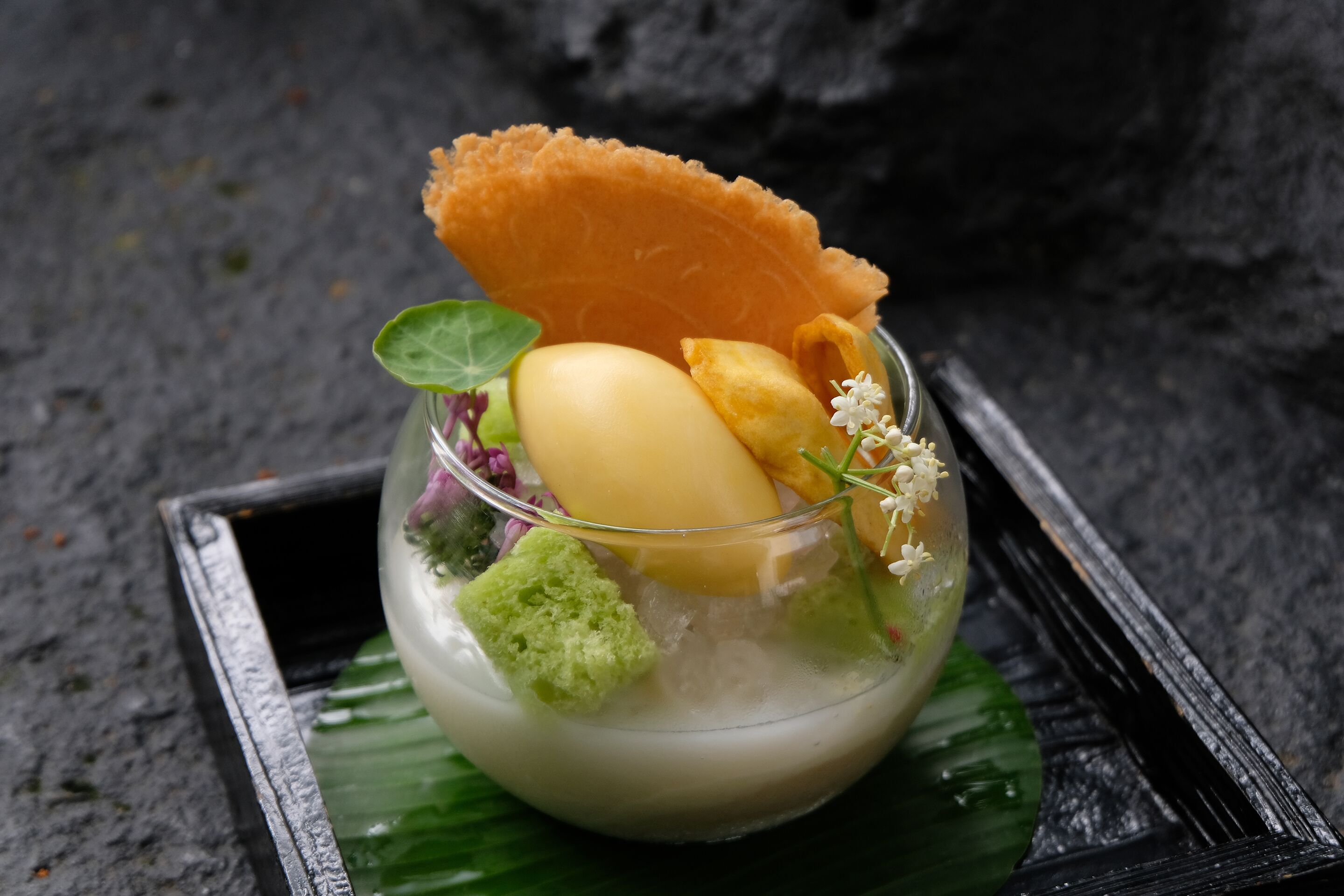 A dessert with a glass bowl, a banana, a green sponge, and a decorative leaf on a black tray.