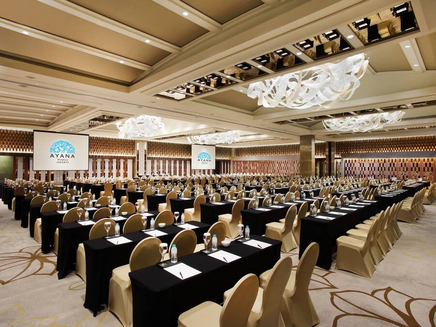 A conference room with rows of tables and chairs, a large screen, and a logo on the wall.