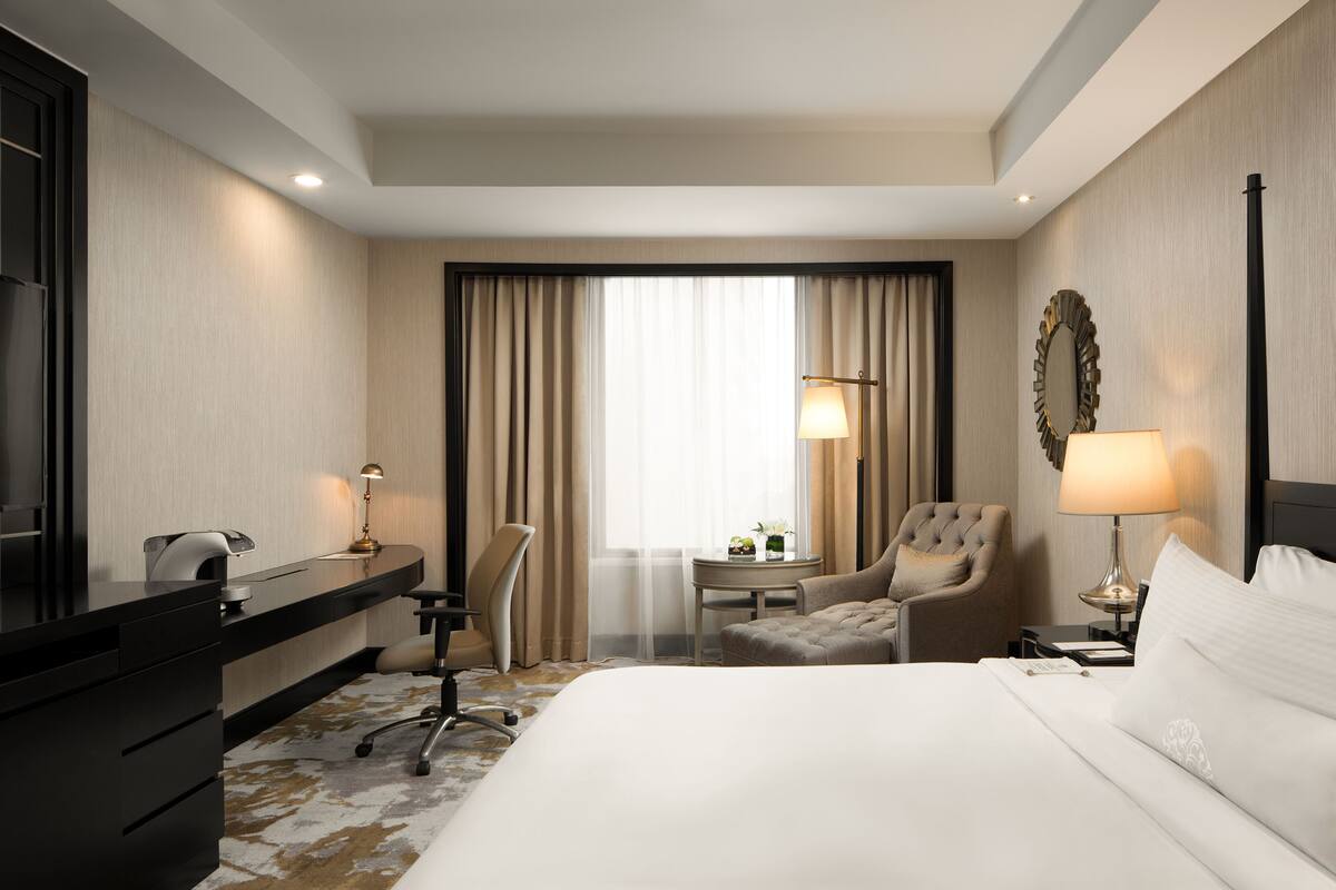 A hotel room with a large bed, a desk with a chair, a small table with a lamp, and a large mirror on the wall.