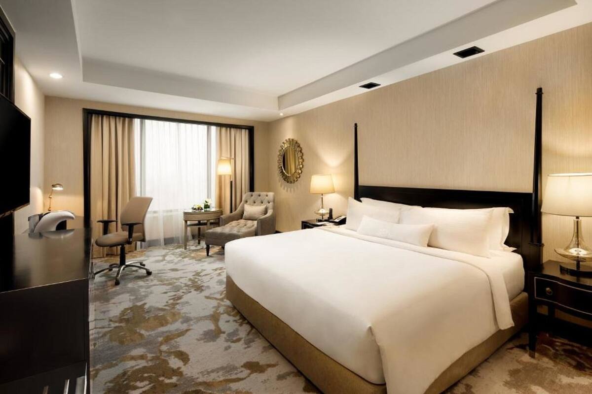 Una habitación de hotel con una cama grande, un escritorio, una silla, un televisor y un ventanal con cortinas.