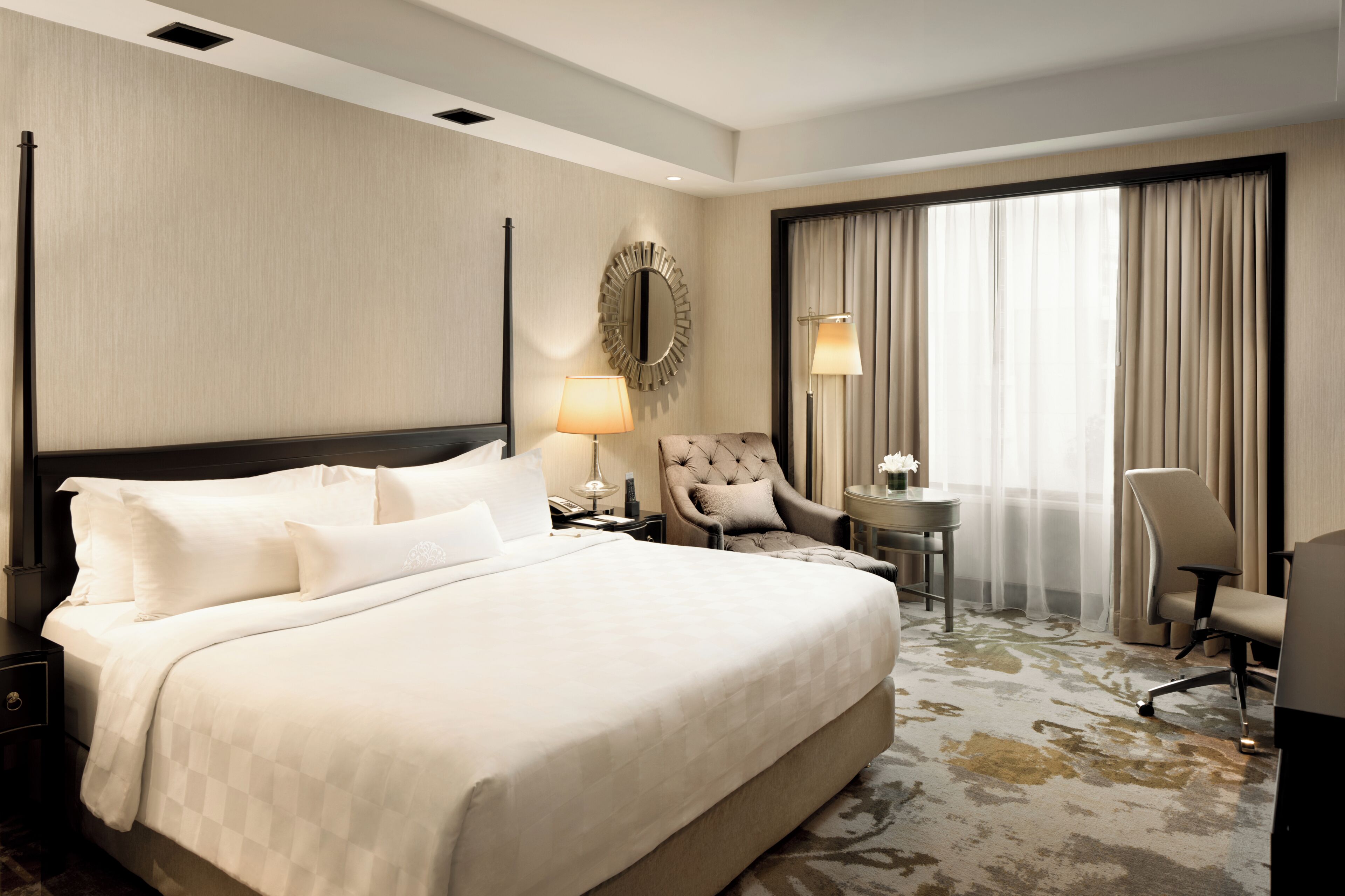 Una habitación de hotel con una cama grande, dos sillones, un espejo redondo, una mesita con una lámpara y un ventanal con cortinas.
