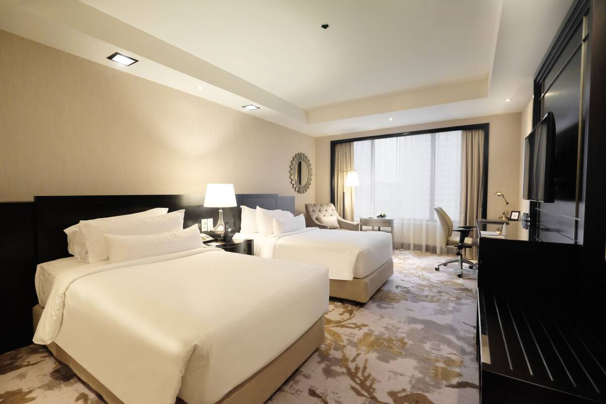 A hotel room with two beds, a desk, a chair, a TV, and a large window.
