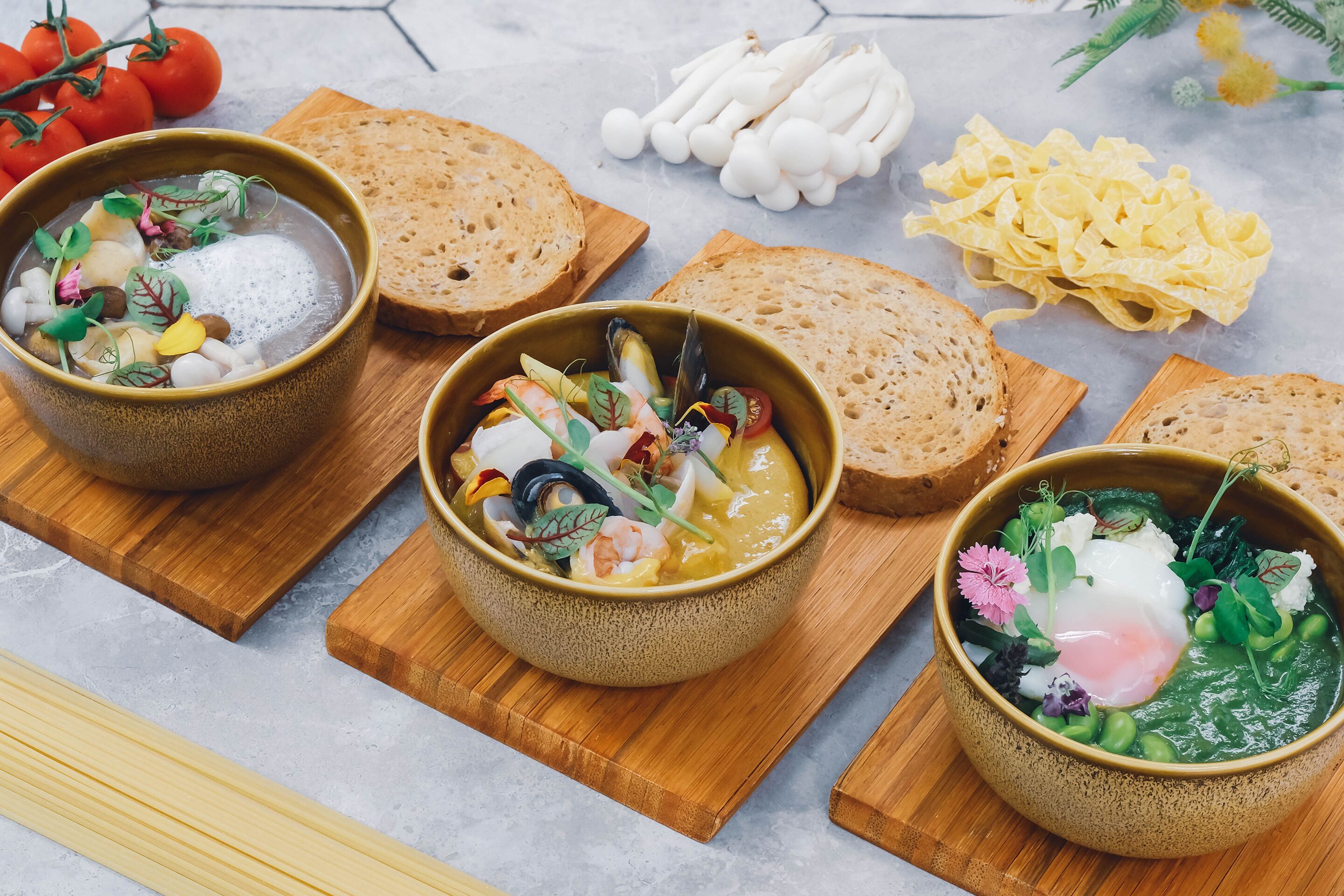Three bowls of soup with various vegetables and garnishes, accompanied by slices of bread and pasta on wooden boards.