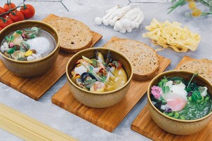 Three bowls of soup with various vegetables and garnishes, accompanied by slices of bread and pasta on wooden boards.