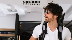 A person in a white t-shirt and black pants with a backpack, standing in a modern interior with a bar area and stools. - Cocoon City Hostel (Chania)