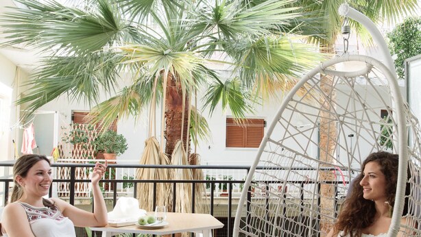 Two people sitting in a hanging chair and on a stool in a courtyard with palm trees and a white table. - Cocoon City Hostel (Chania)